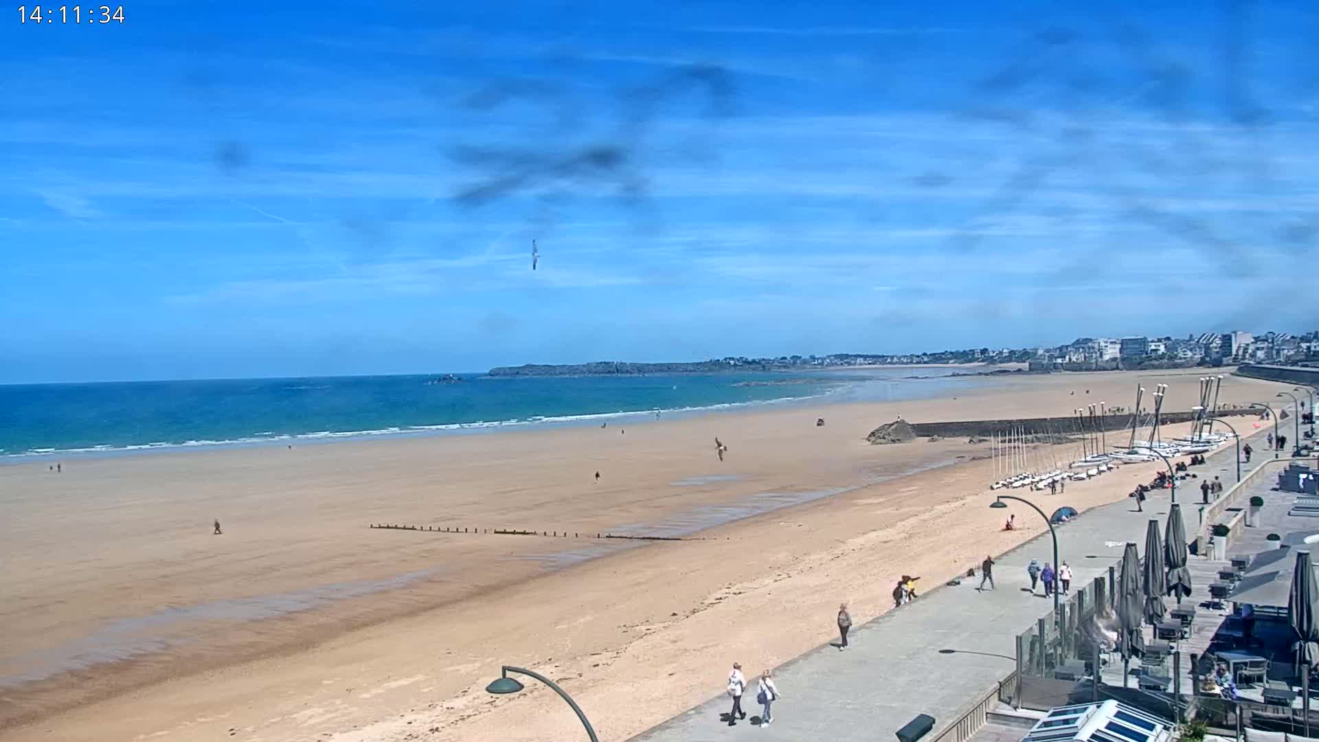 A sandy beach stretches along a calm ocean under a mostly sunny sky, with people walking along a paved walkway near a line of buildings.