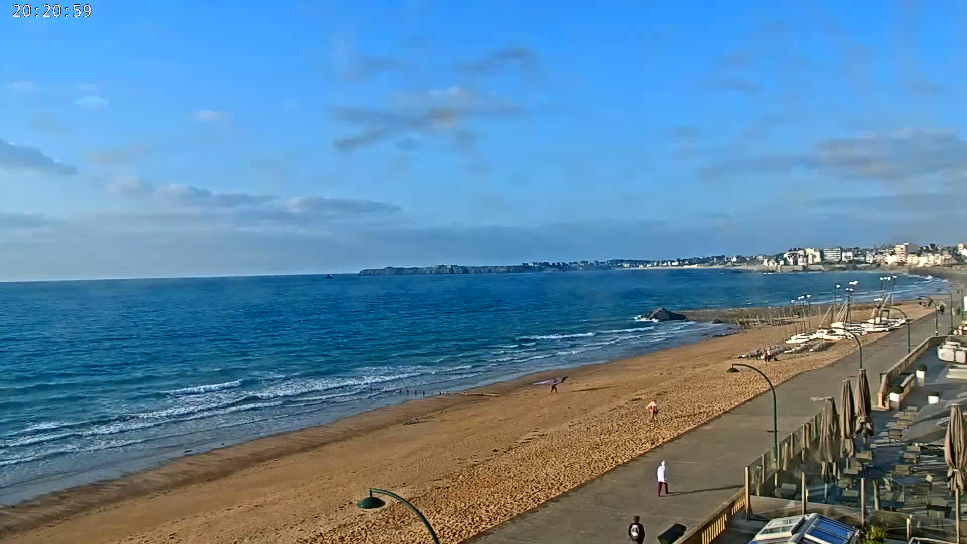 A sandy beach with gentle waves under a mostly clear blue sky stretches to a distant town, with a few people strolling along the promenade.