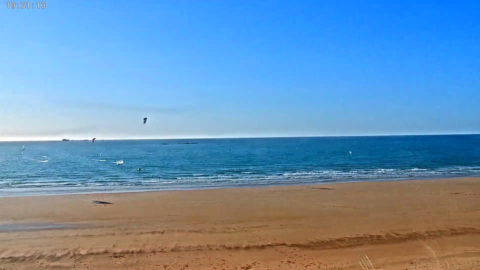 A sandy beach stretches to a calm, blue ocean where several kitesurfers are enjoying a sunny day.