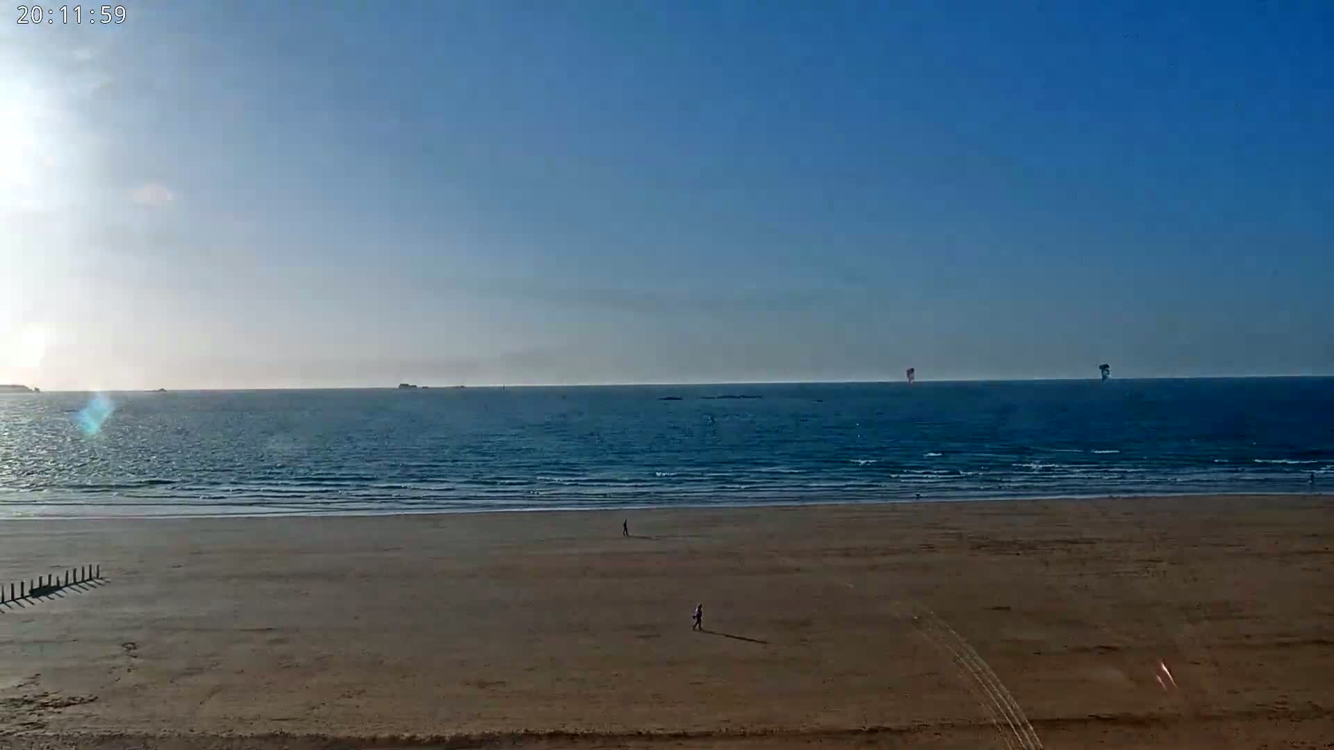 A mostly clear, sunny day shows a wide expanse of sandy beach and calm ocean with a few people walking on the sand.