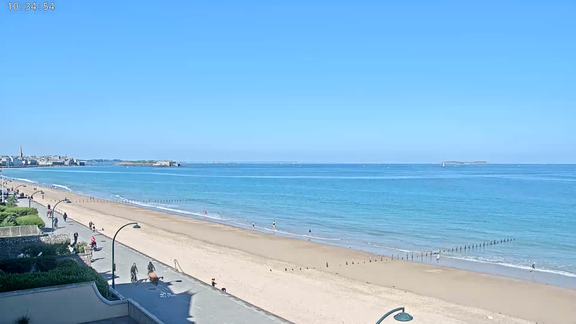A sandy beach with people walking and biking along a paved path stretches towards a calm, light blue ocean under a clear blue sky.