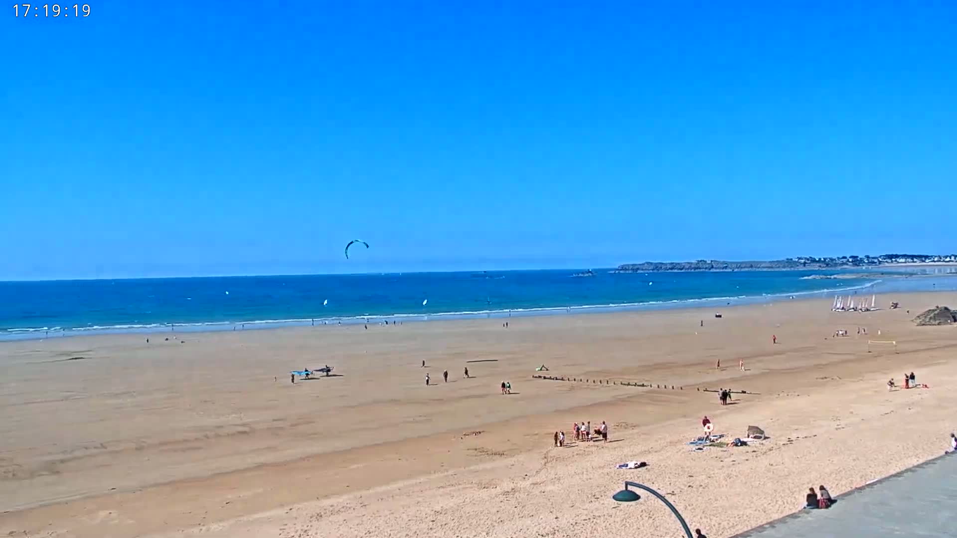 A wide sandy beach stretches to a calm ocean under a clear blue sky, with people enjoying the sunshine and several kitesurfers and windsurfers in the water.