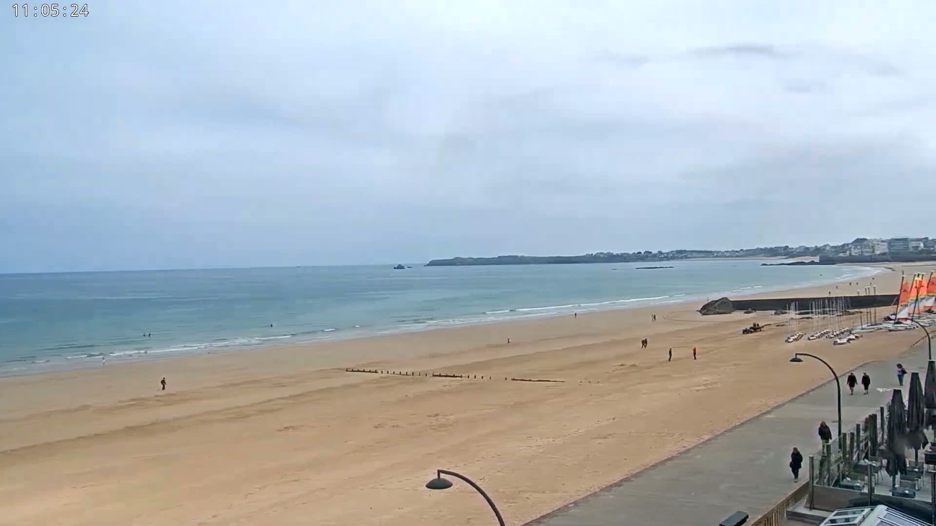 A wide sandy beach stretches to a calm ocean under an overcast sky, with a few people scattered on the sand and a row of sailboats parked near a boardwalk.