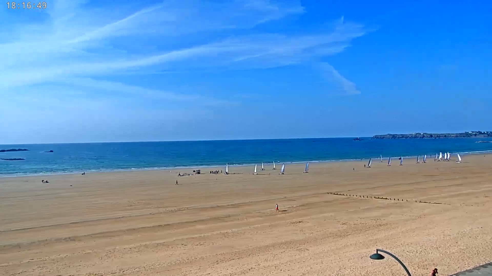 A mostly sunny day at a beach shows a wide expanse of sand with several small sailboats and a few people scattered about, and a calm ocean beyond.