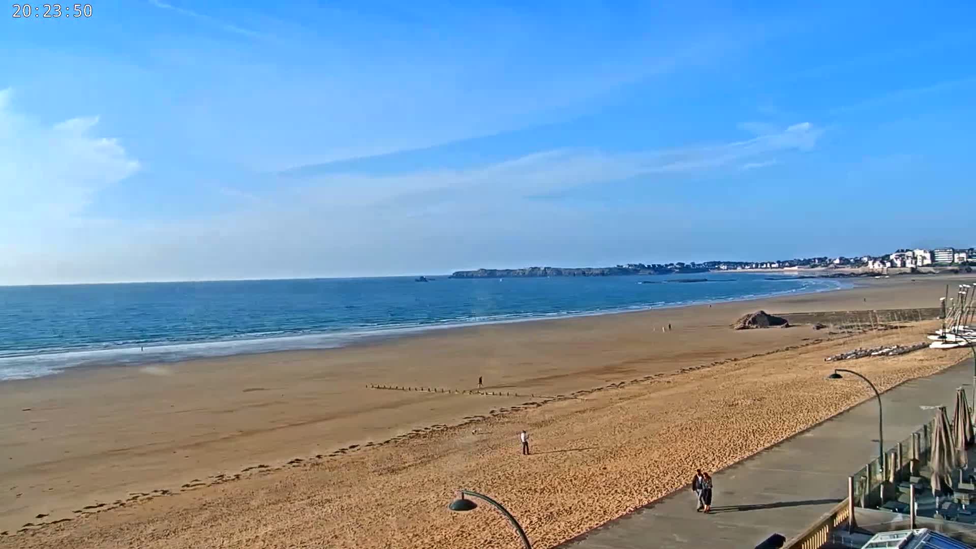 A wide sandy beach stretches to a calm ocean under a mostly clear blue sky, with a few people walking along the shore and a paved walkway nearby.
