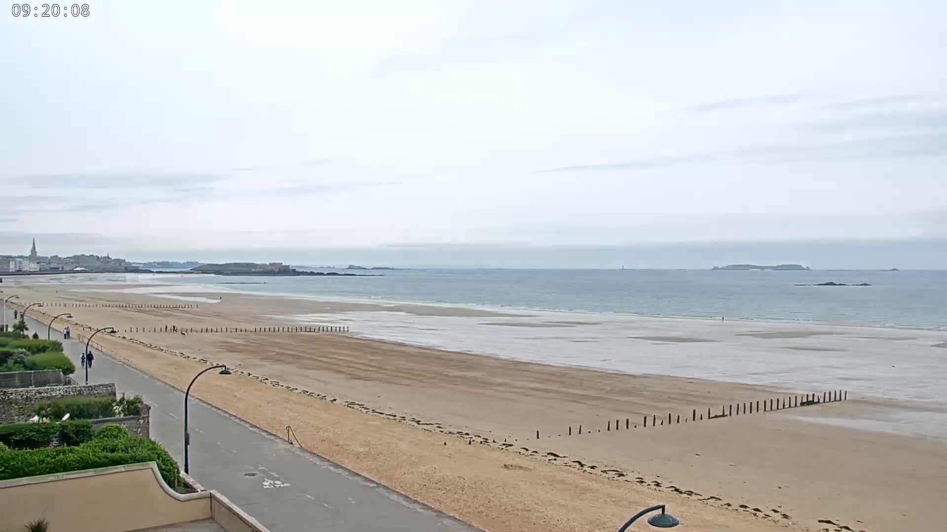 A wide sandy beach stretches out to a calm, light-blue ocean under an overcast sky, with a paved walkway and vegetation bordering the beach.