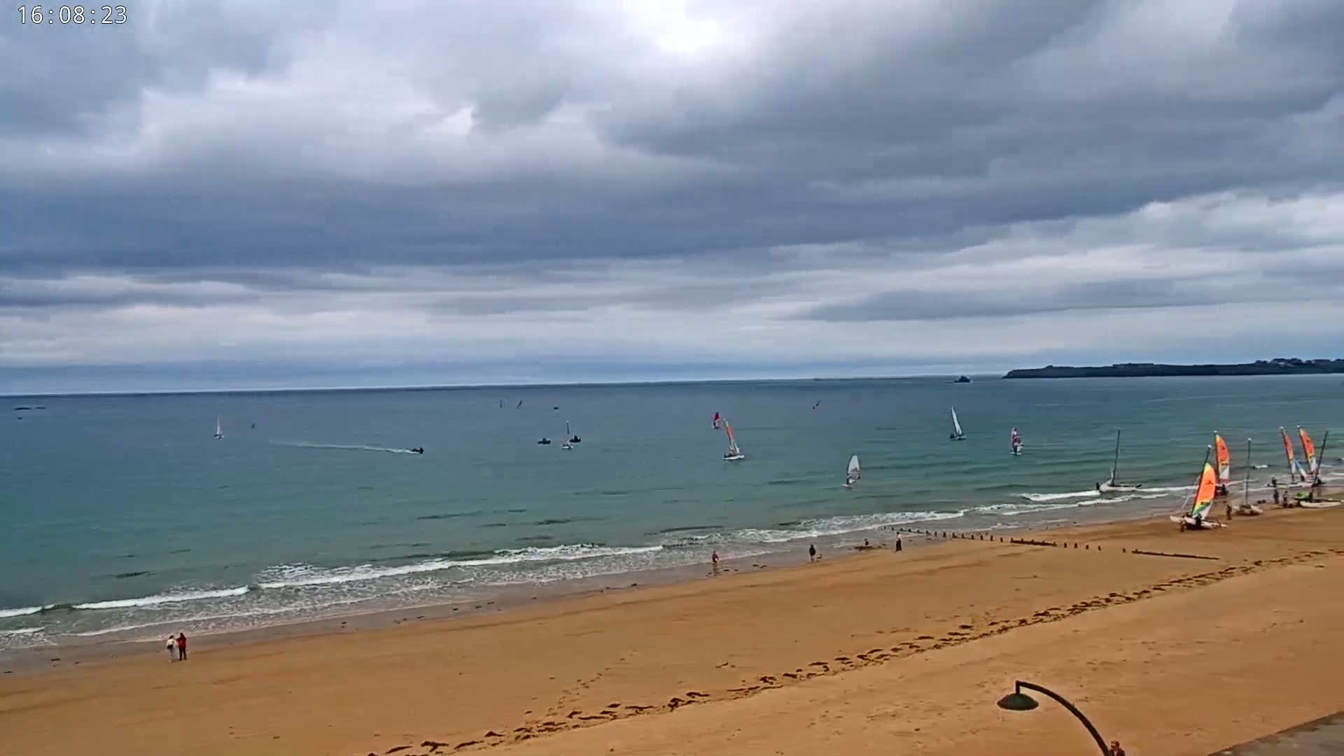 A sandy beach with several sailboats in the ocean under a cloudy sky.