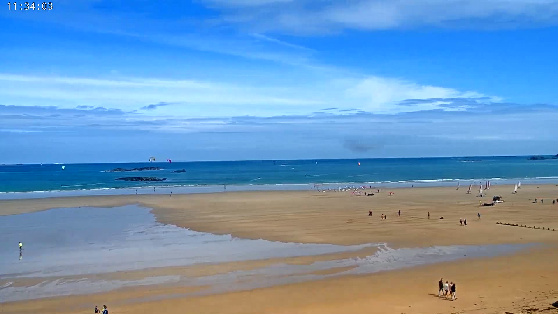 A sandy beach with scattered people and several sailboats and kitesurfers in the water under a mostly sunny sky.