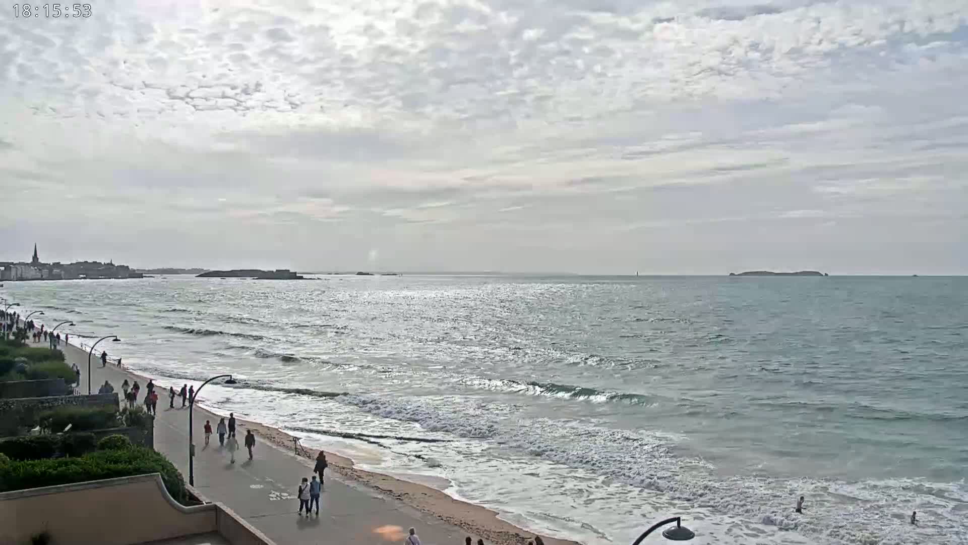 A mostly cloudy day shows a beach with people walking along a promenade, gentle waves lapping the shore, and a town visible in the distance across the water.