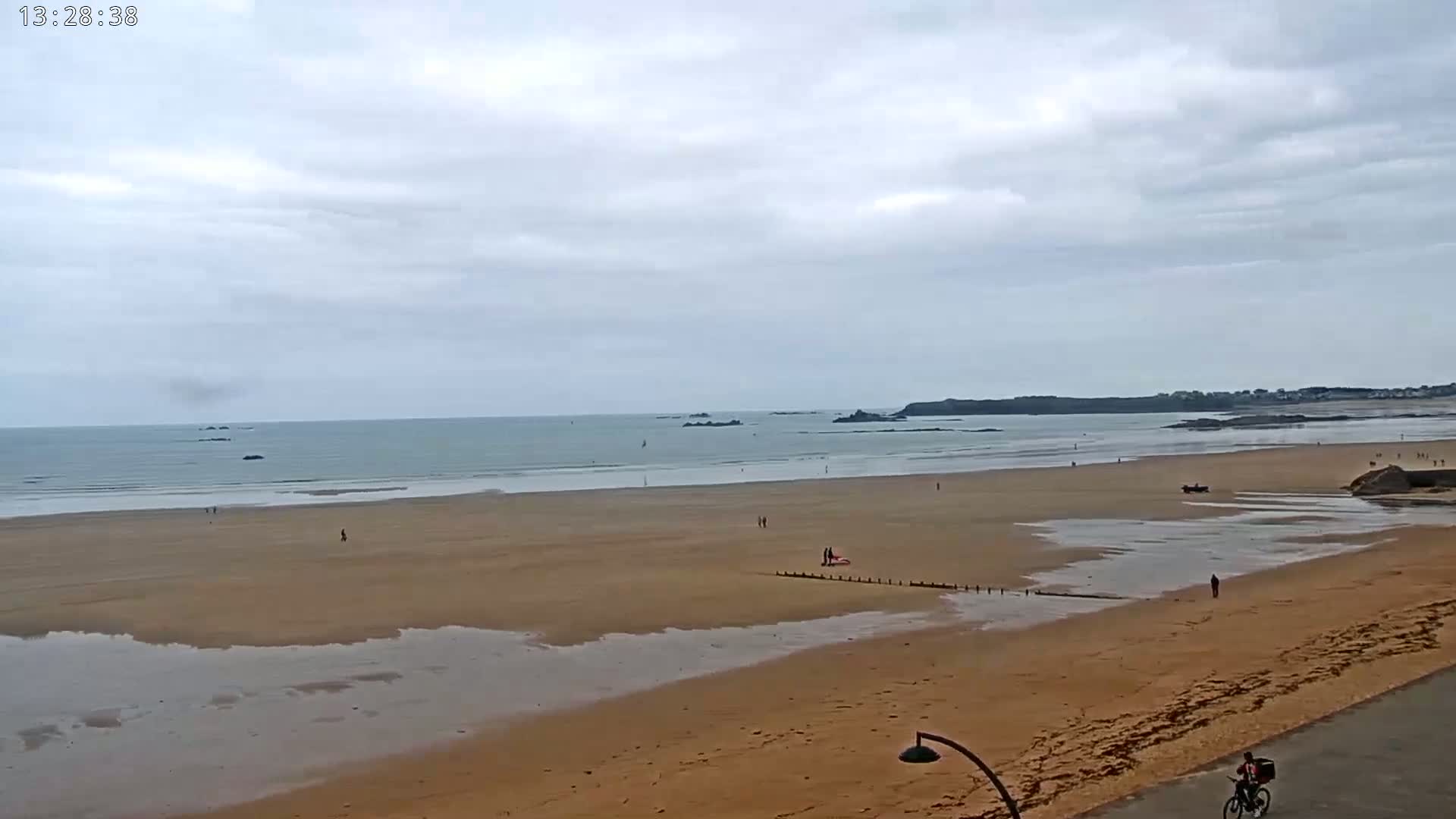 A wide sandy beach stretches to a calm ocean under a cloudy sky, with a few people scattered across the sand and a lone cyclist on a nearby road.