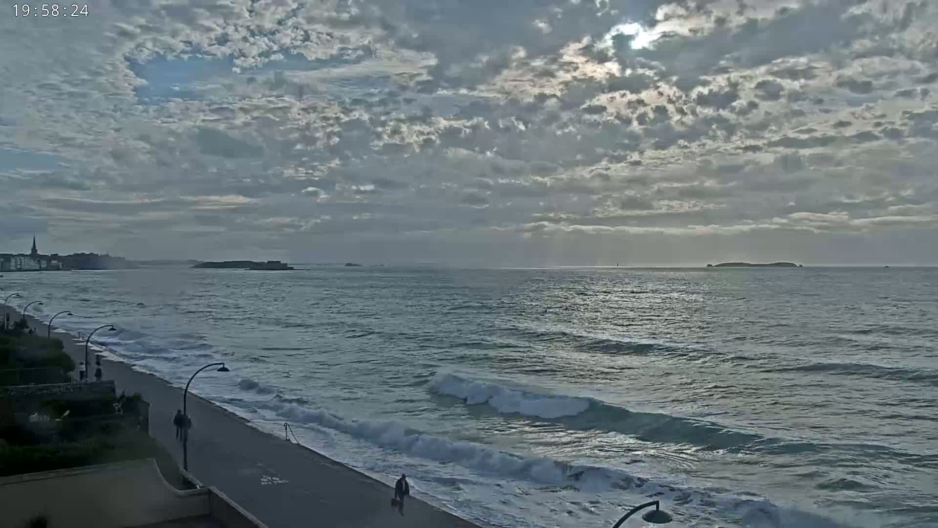 A mostly cloudy sky over a wavy ocean with a paved beachfront walkway and a few people walking.