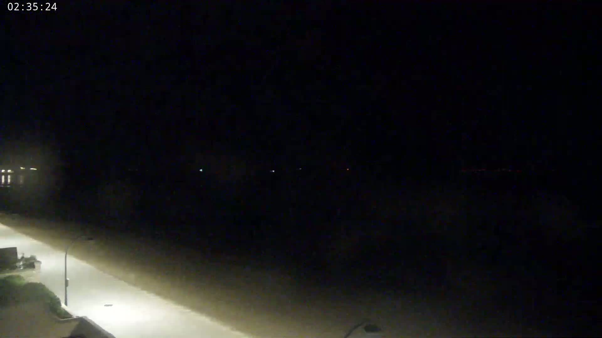 A nighttime, dimly lit view shows a paved road extending toward a dark body of water under a clear sky.