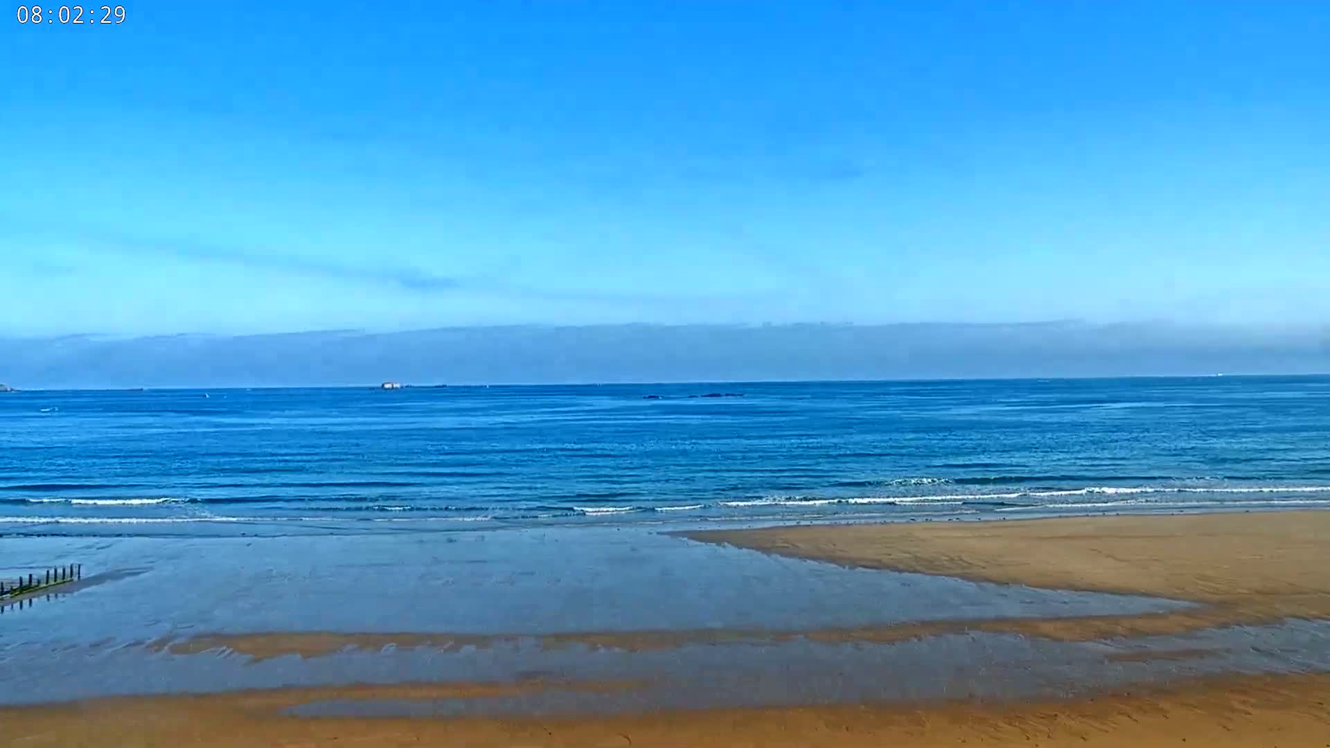 A mostly sunny day at a beach shows calm, blue water lapping a wide, sandy shore.