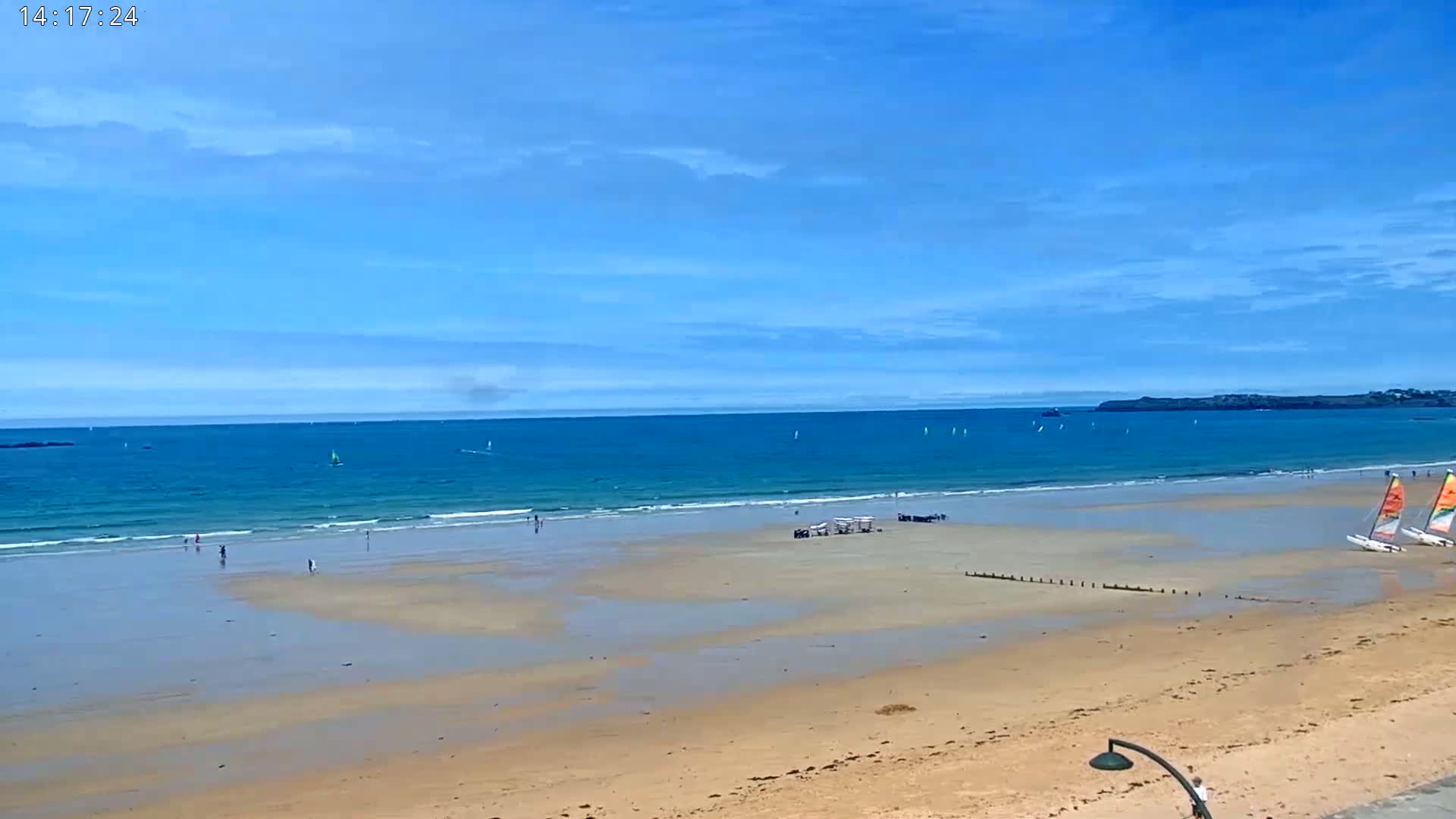 A wide sandy beach stretches to a calm, blue ocean under a mostly clear sky, with a few sailboats visible in the water and people scattered along the shore.
