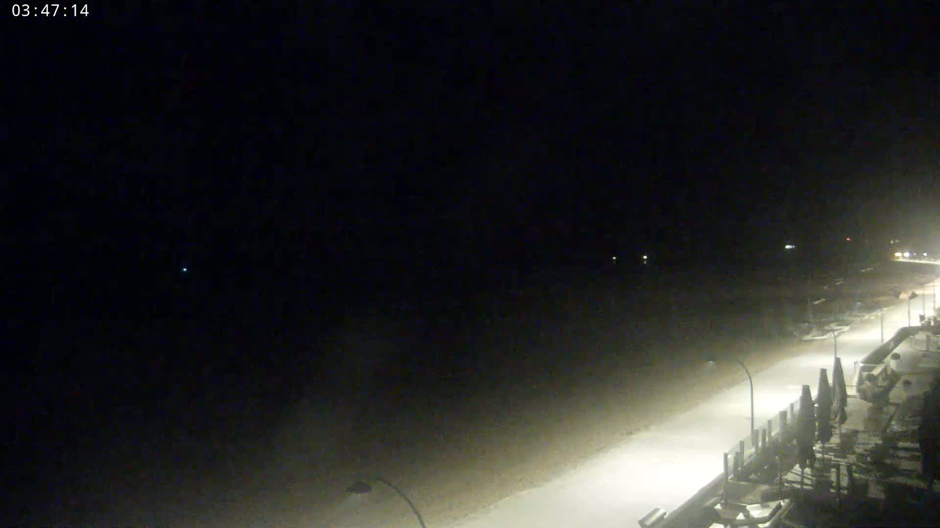 A snow-covered beach at night is illuminated by streetlights, with some objects, possibly beach umbrellas, visible in the lower right corner.