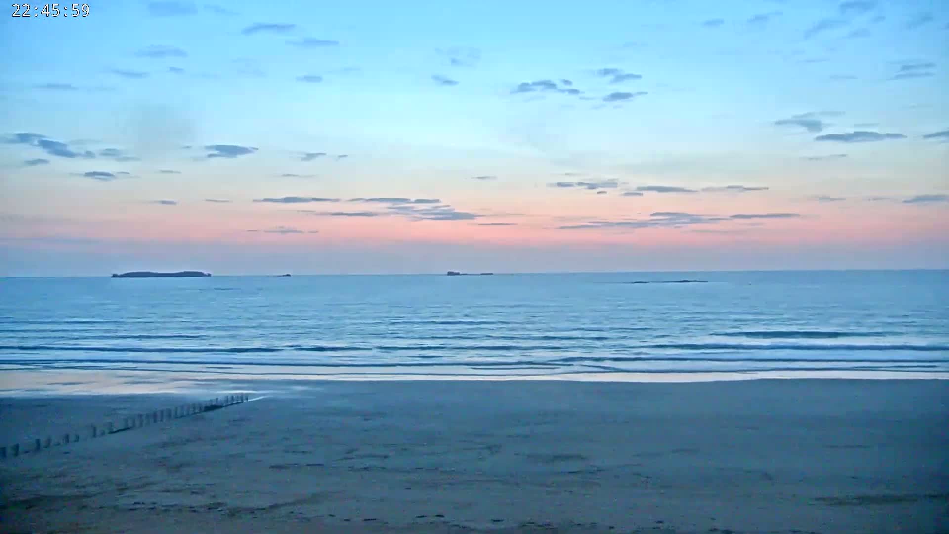 A calm ocean with small waves washes a sandy beach under a pastel sunset sky.