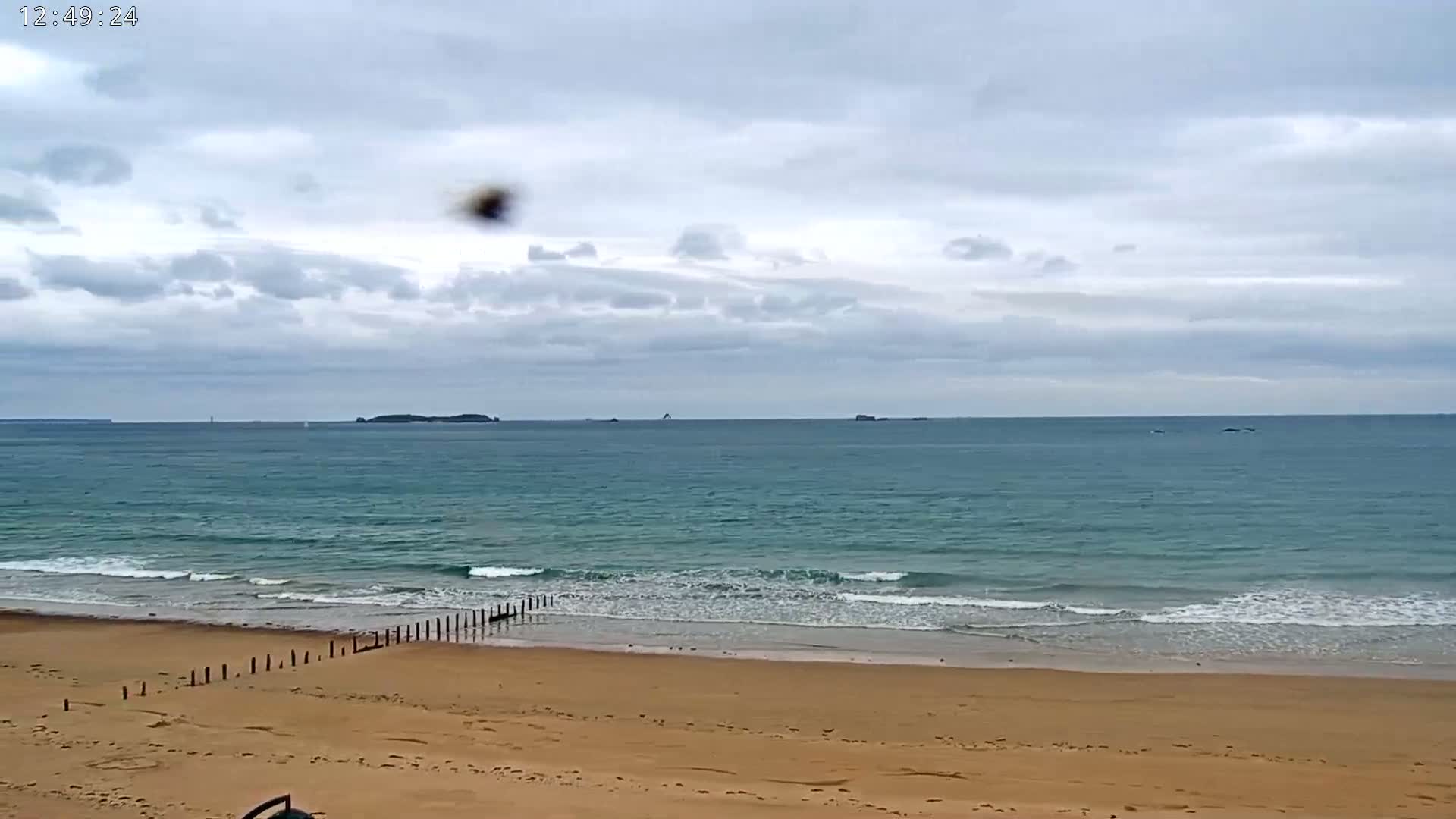 A sandy beach with small waves laps the shore under a mostly cloudy sky, with islands visible on the horizon.
