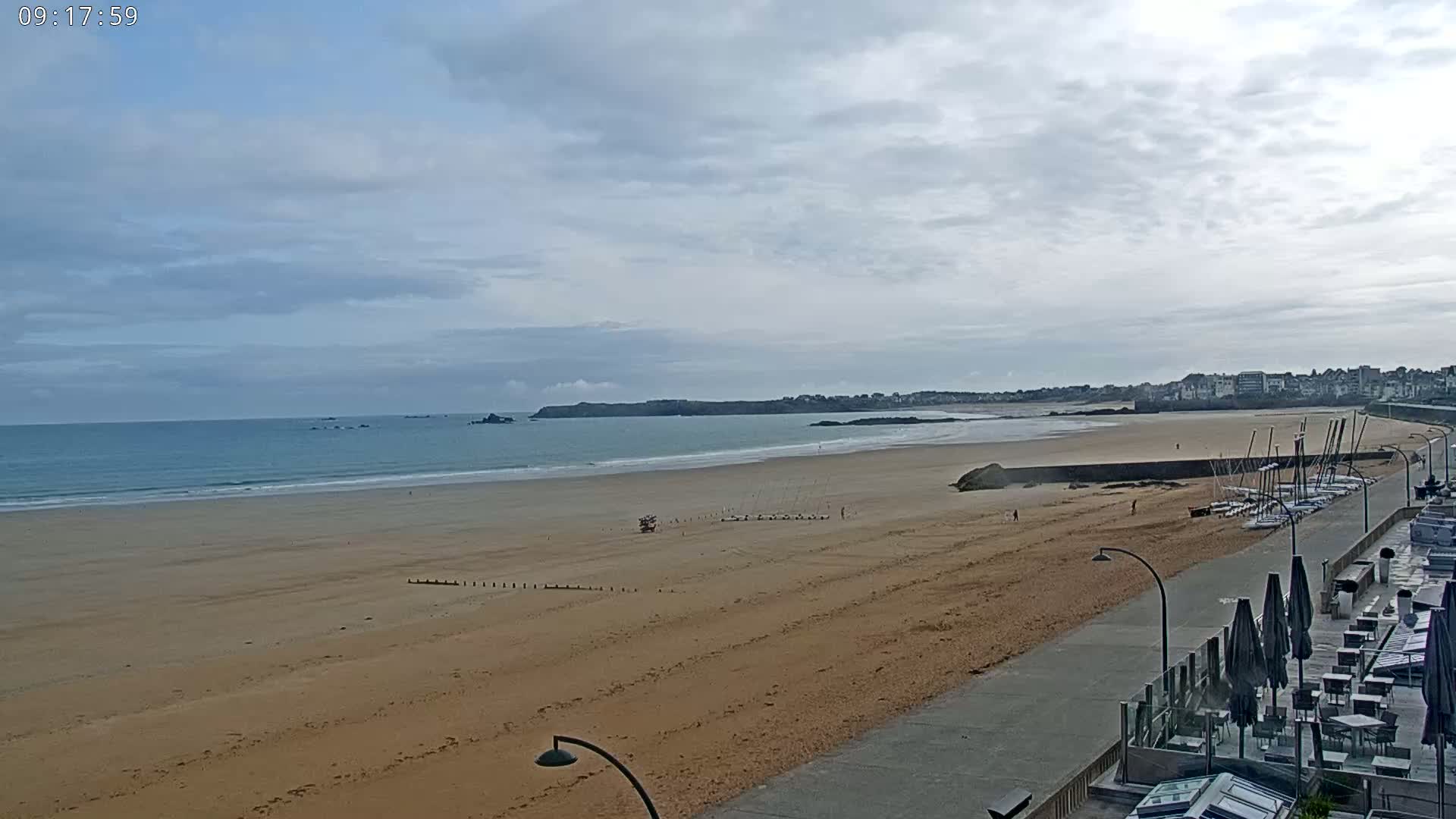 A wide sandy beach stretches to a calm ocean under a mostly cloudy sky, with a paved walkway and outdoor seating area visible in the foreground.