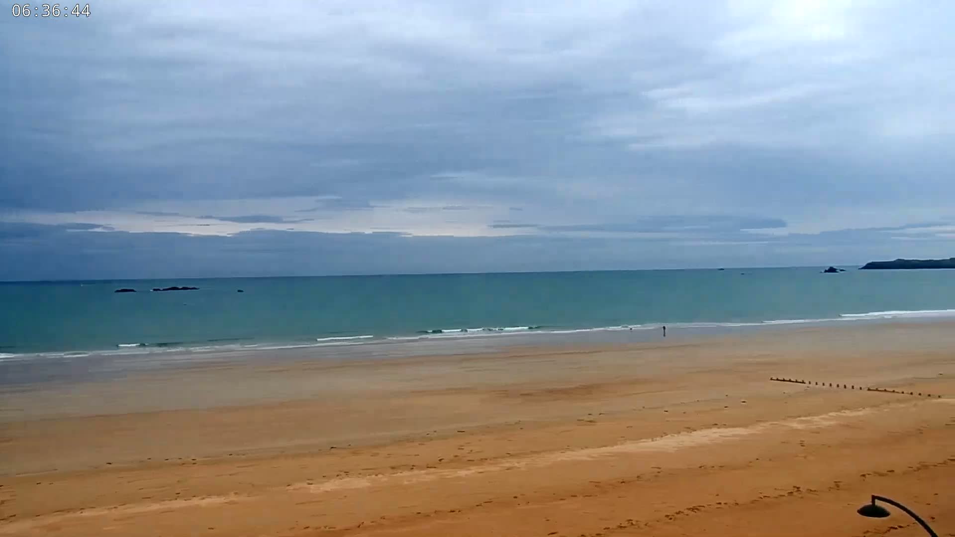 A wide expanse of sandy beach meets a calm, teal ocean under a mostly cloudy sky.