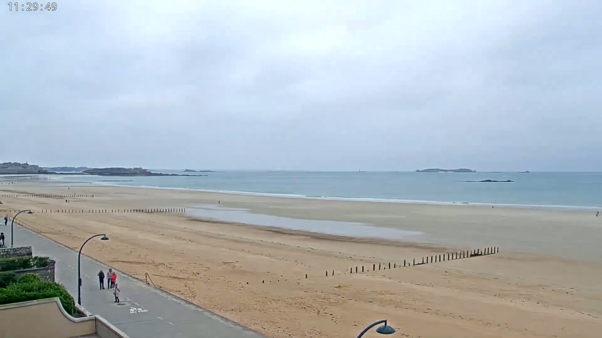 A long, sandy beach stretches to a calm ocean under an overcast sky, with a few people strolling along the shore and a paved walkway nearby.
