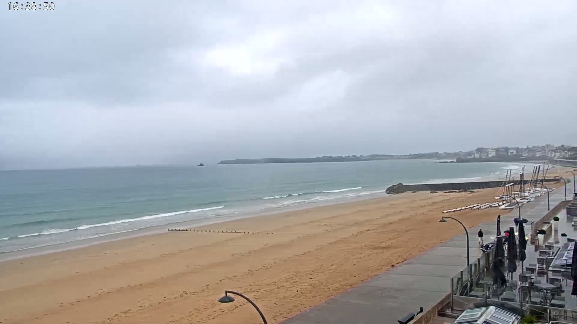 A sandy beach with gentle waves stretches along a coastline under a cloudy sky, with sailboats visible near a building and a paved walkway.