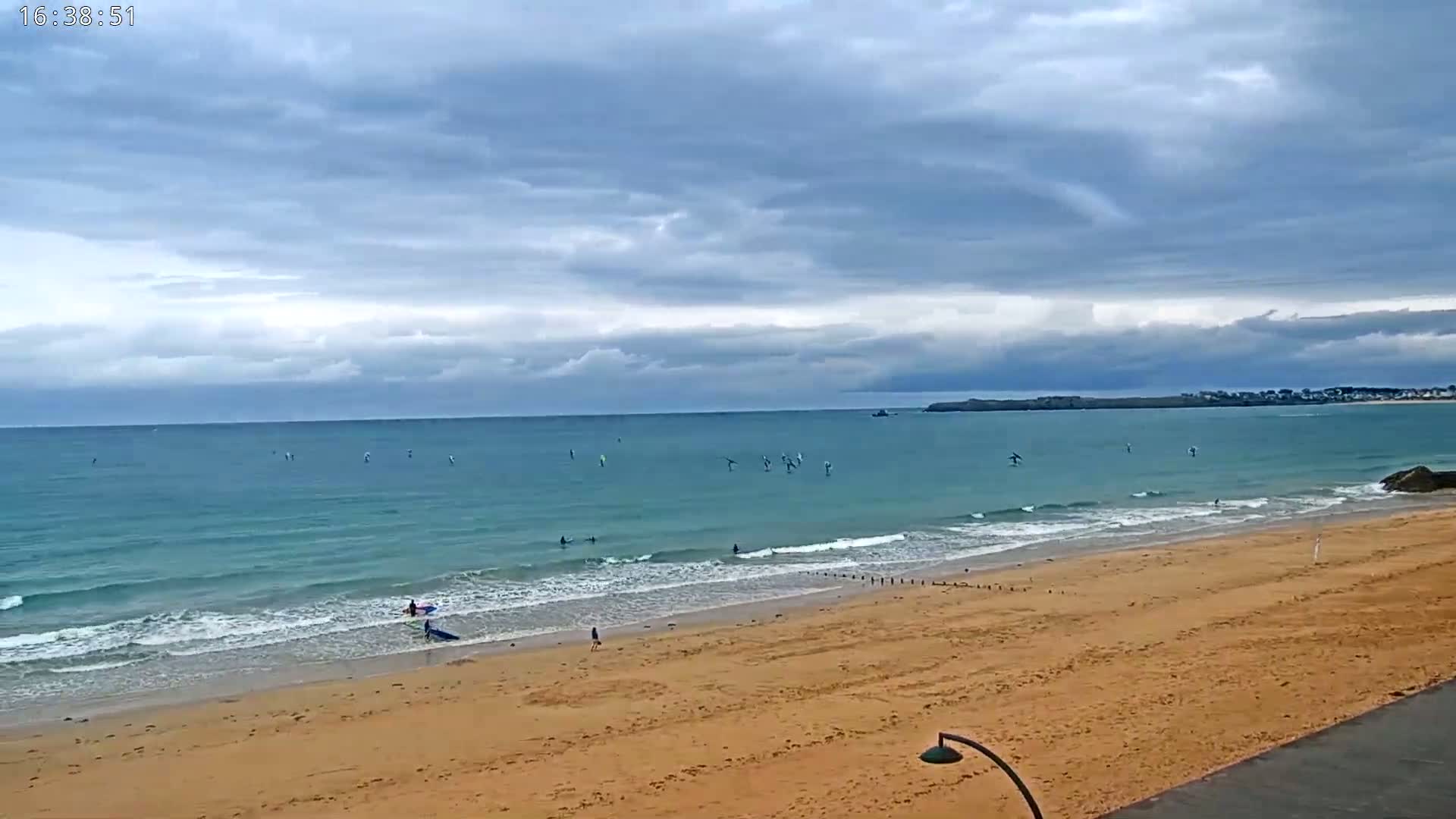 A sandy beach with several people and windsurfers on the water under a cloudy sky.