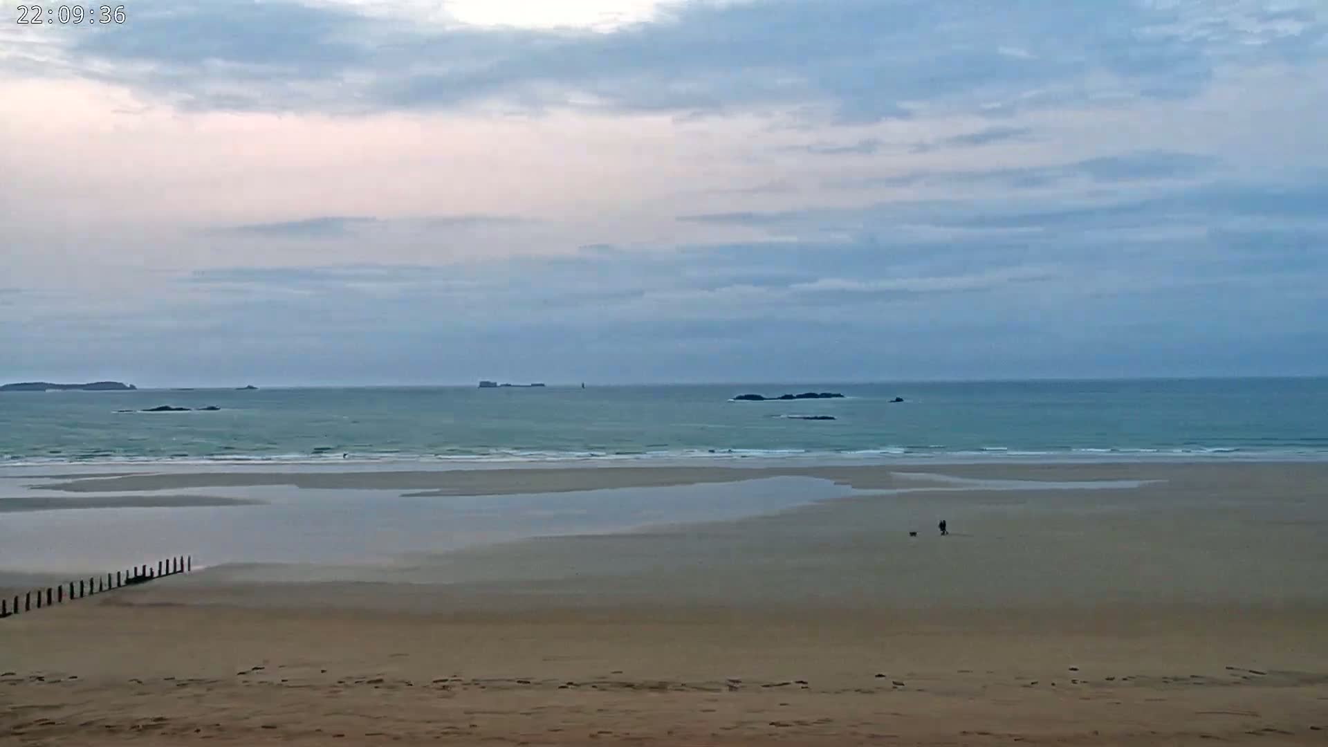 A sandy beach stretches out to a calm, light teal ocean under a mostly cloudy, pale sky, with two people and a dog walking near the waterline in the distance.