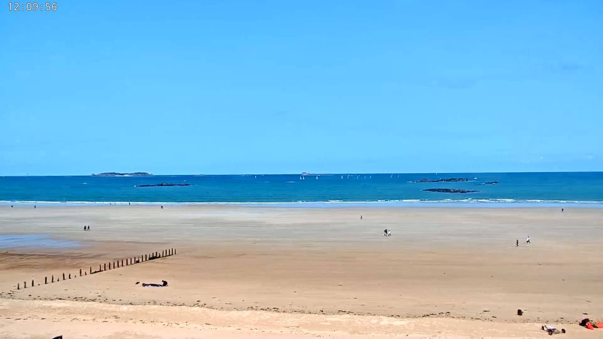 A wide sandy beach stretches toward a calm, turquoise ocean under a clear blue sky, with a few people scattered across the sand and several sailboats visible on the horizon.