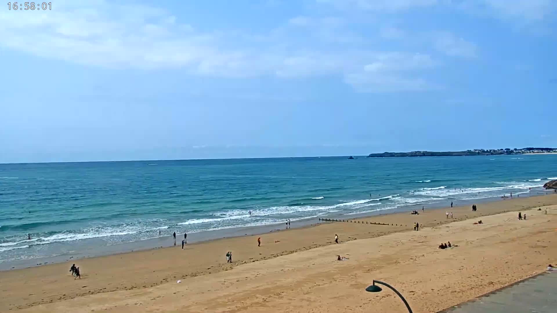 A sandy beach with a few people on it slopes down to a calm, teal ocean under a mostly clear, bright blue sky.