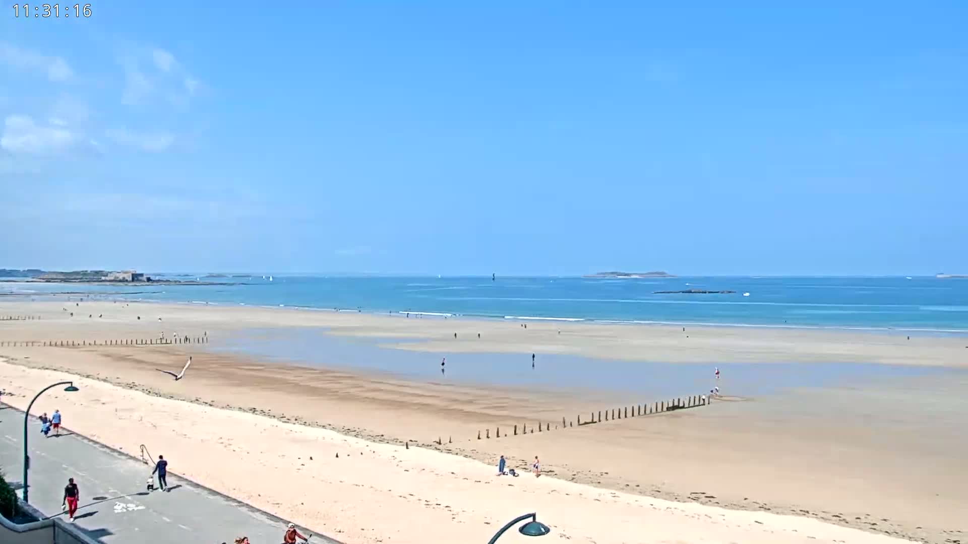 A wide sandy beach with people scattered across it and a calm ocean under a mostly clear blue sky.