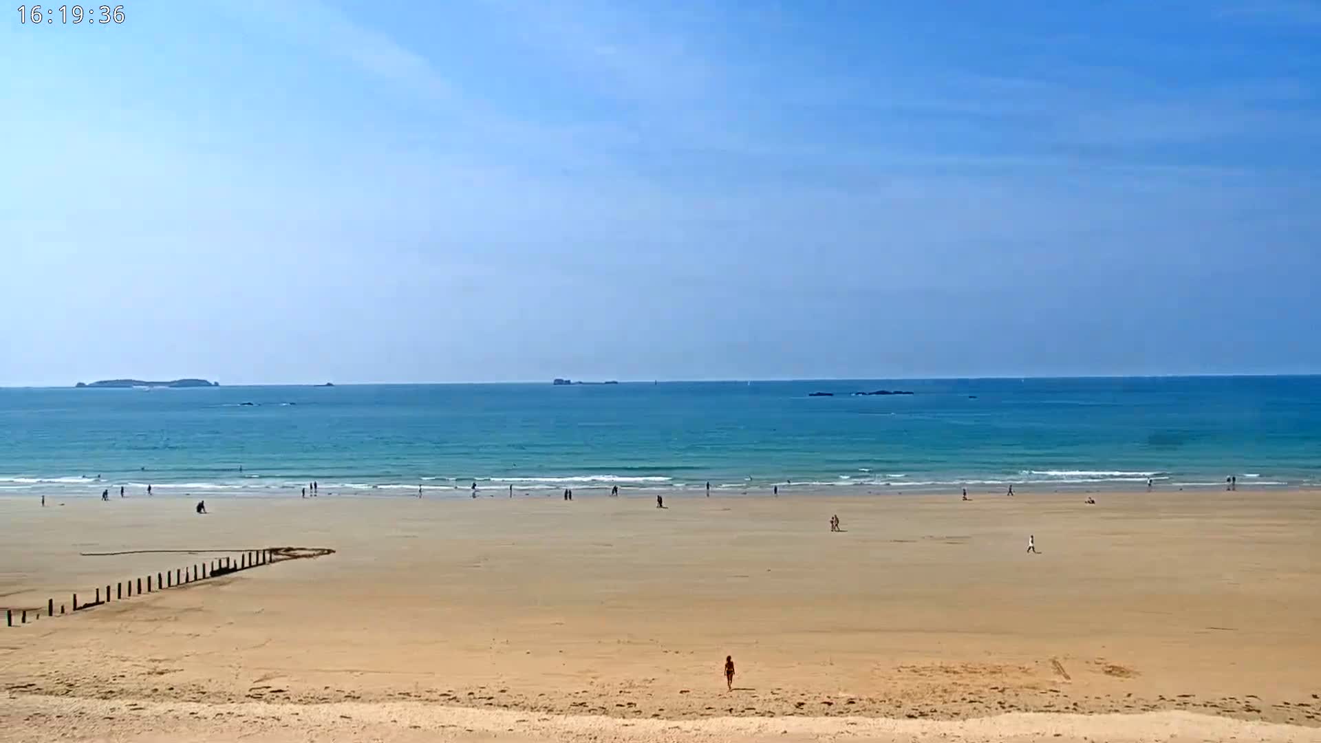 A sandy beach stretches to a calm, turquoise ocean under a clear blue sky, with a few people scattered across the sand and in the water.
