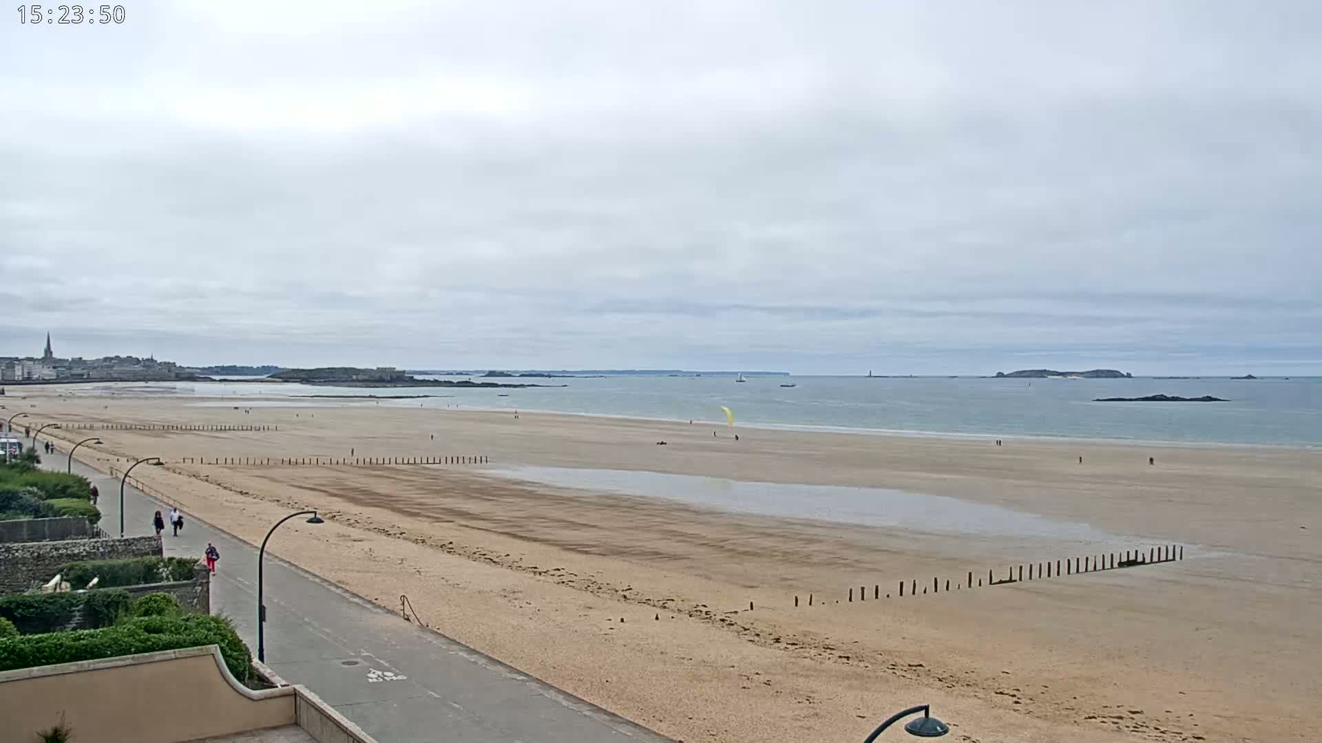 A wide sandy beach stretches to a calm sea under an overcast sky, with a paved walkway and a few people visible.
