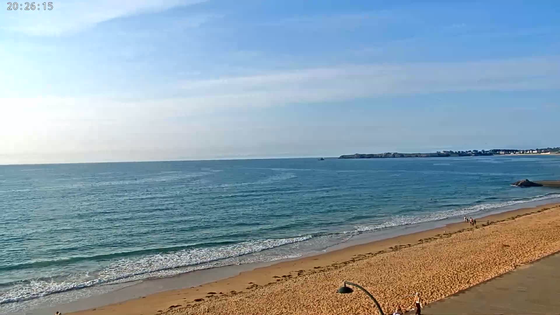 A sandy beach with gentle waves laps the shore under a mostly clear sky, with a few people visible on the sand and in the water.