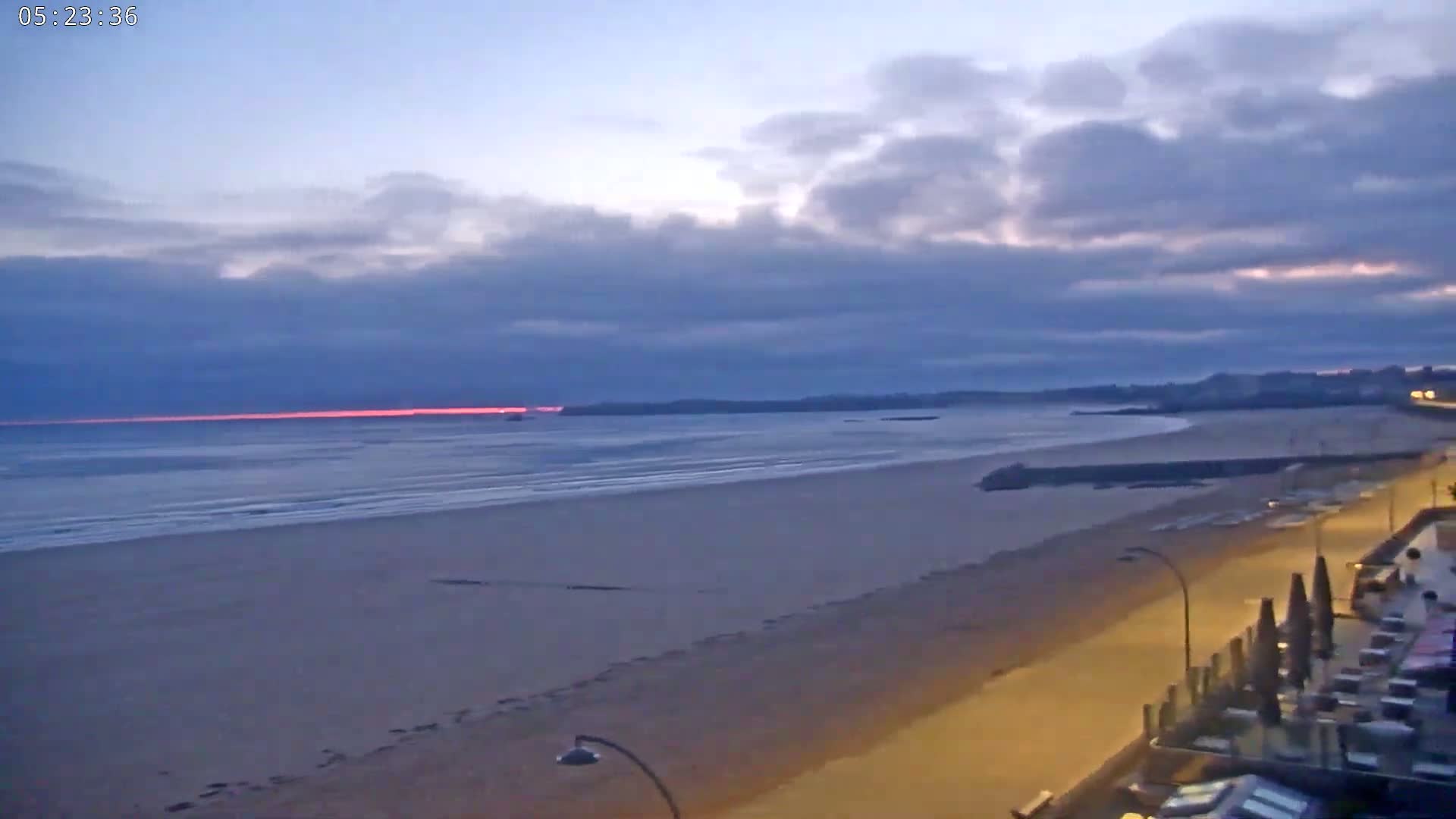 A wide sandy beach stretches to a calm ocean under a cloudy twilight sky with a faint pinkish sunrise on the horizon.