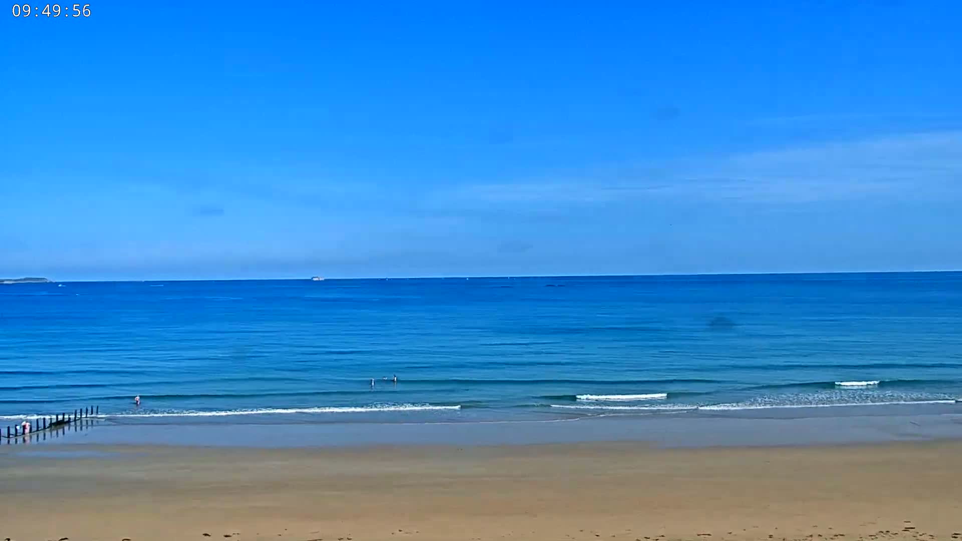 A sandy beach with gently lapping waves meets a calm, blue ocean under a clear, sunny sky.