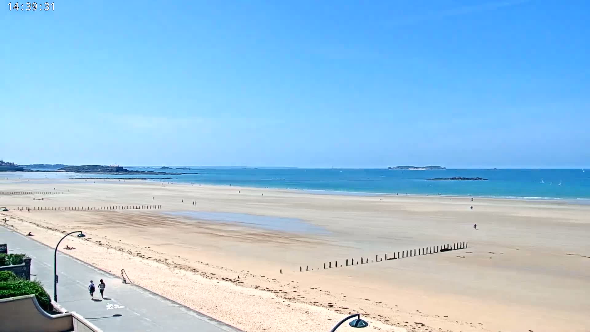A wide sandy beach stretches to a calm, blue ocean under a clear, sunny sky, with a few people scattered across the sand and a paved walkway along the beach's edge.