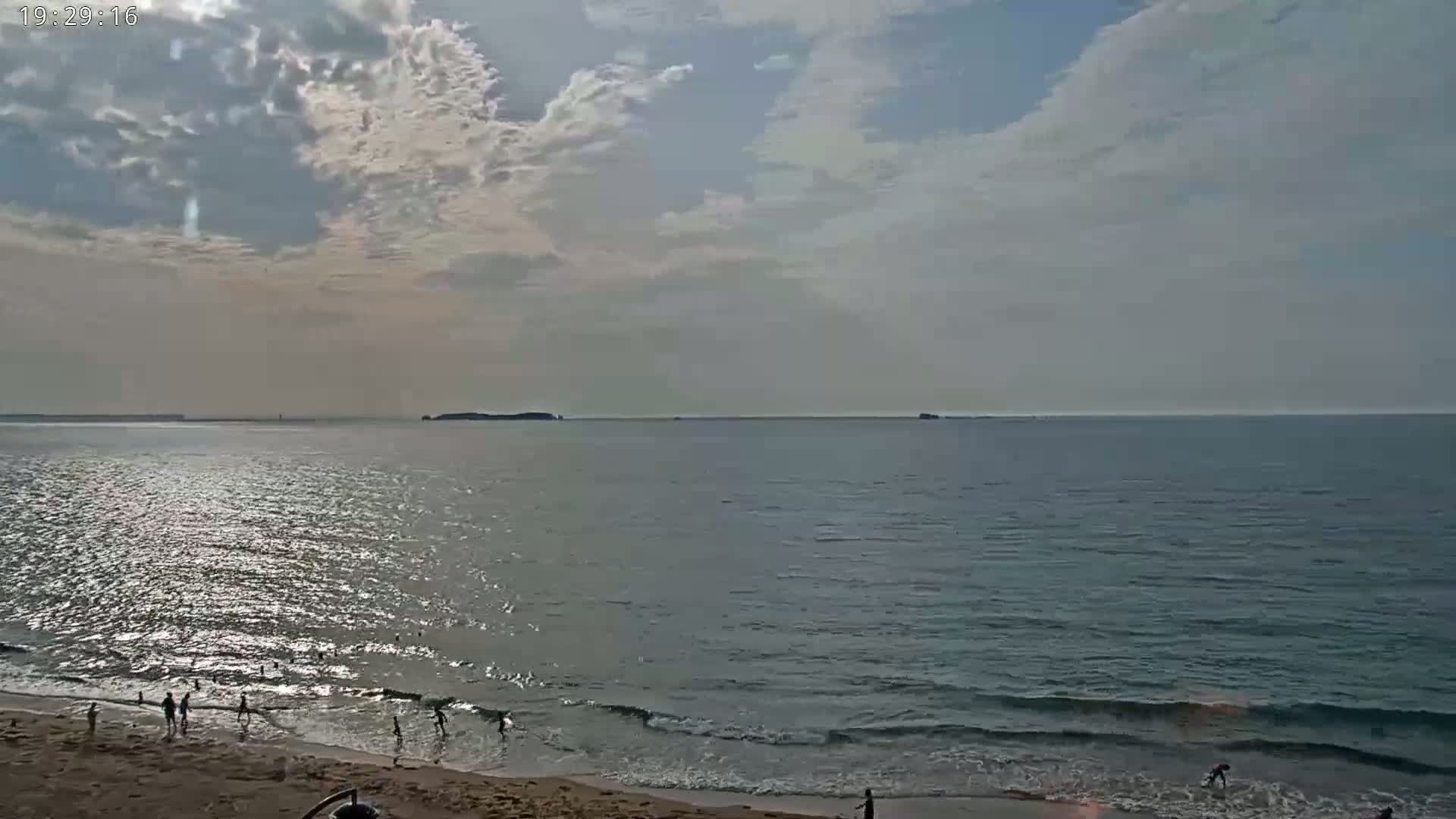 A mostly cloudy sky hangs over a calm ocean beach where several people are wading in the shallows.