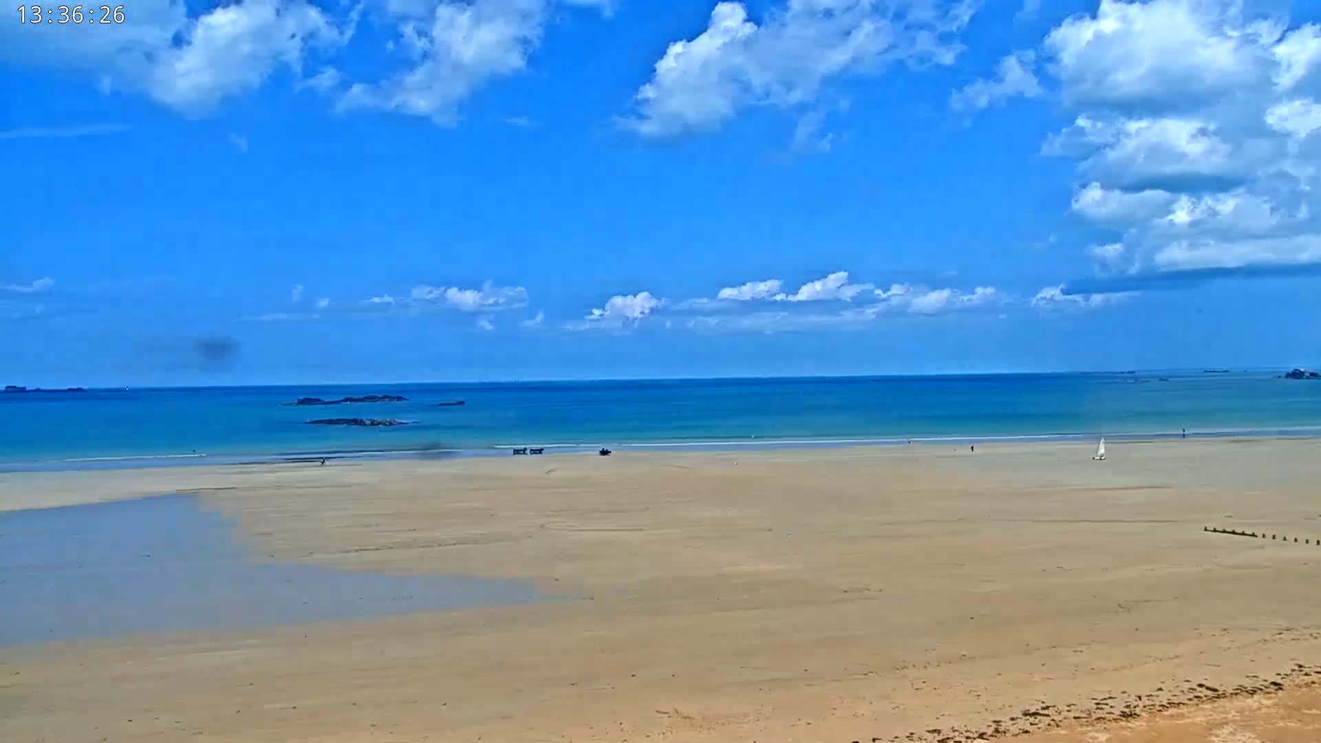 A sandy beach stretches to a calm, turquoise ocean under a mostly sunny sky with scattered puffy clouds.