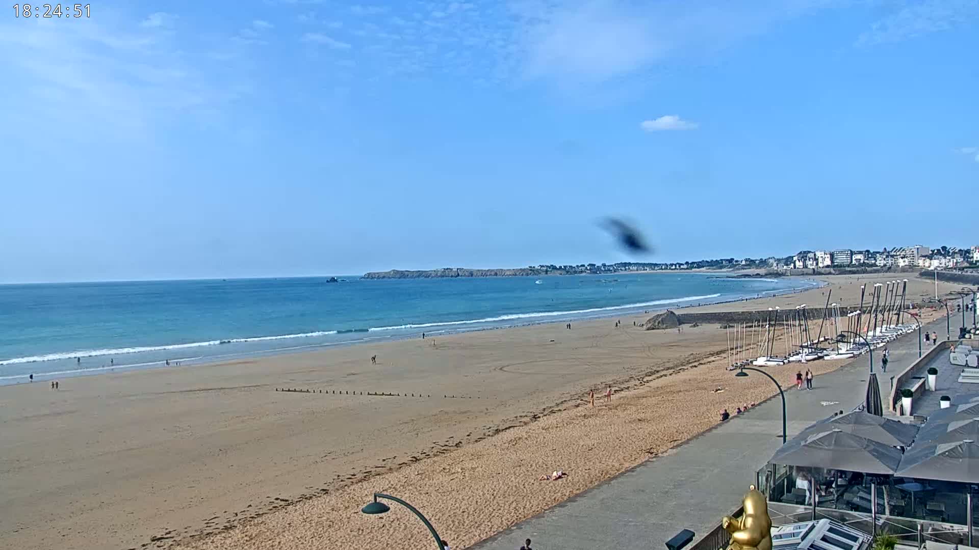 A sandy beach stretches along a calm ocean under a clear blue sky, with small waves breaking near the shore and sailboats beached on the sand.