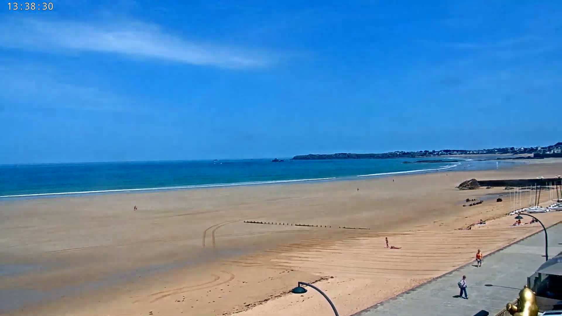 A sandy beach stretches out to a calm, blue ocean under a clear, sunny sky, with a few people scattered along the shore and a town visible in the distance.