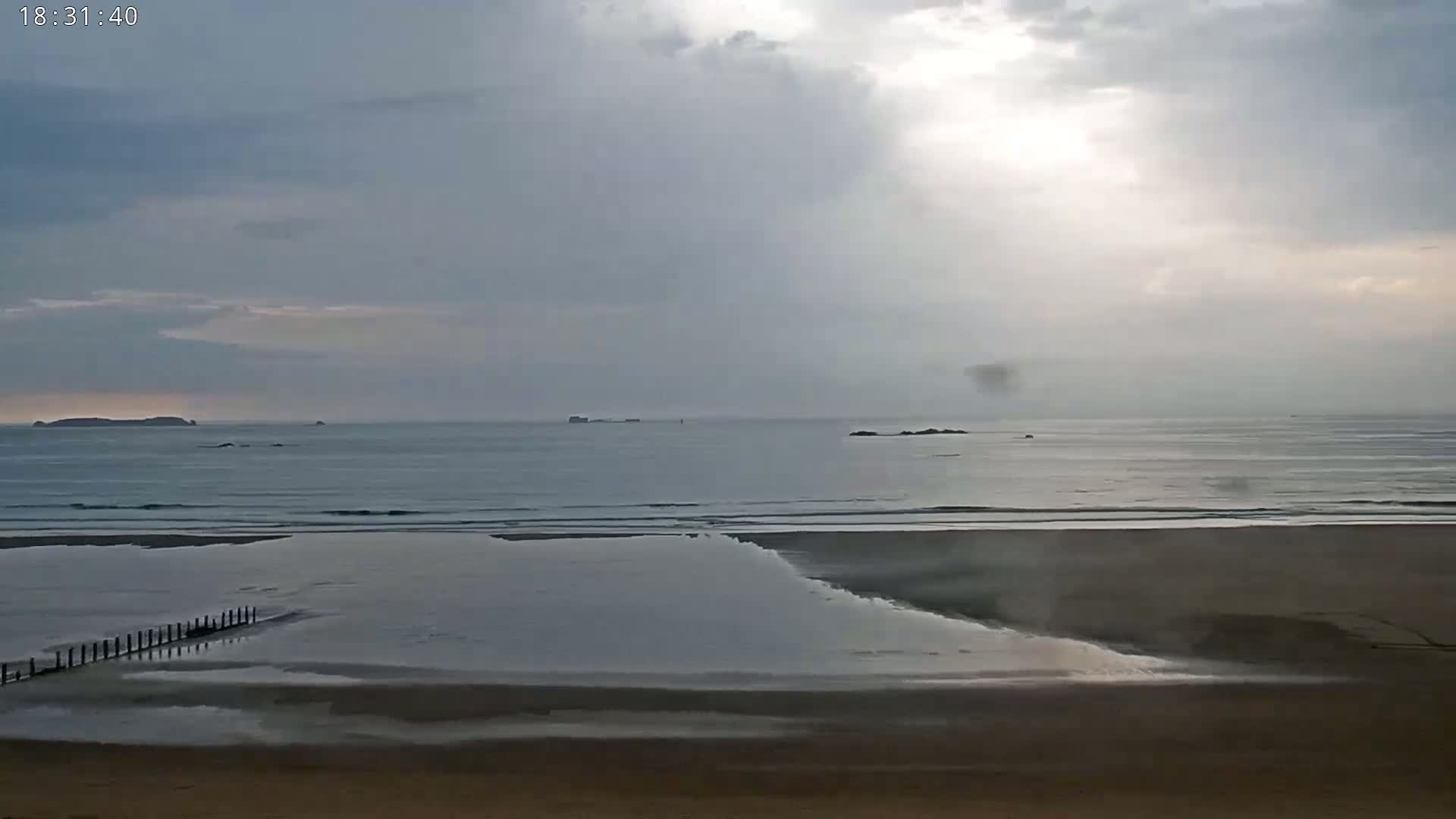 A sandy beach with a calm ocean and overcast skies shows distant islands on the horizon.