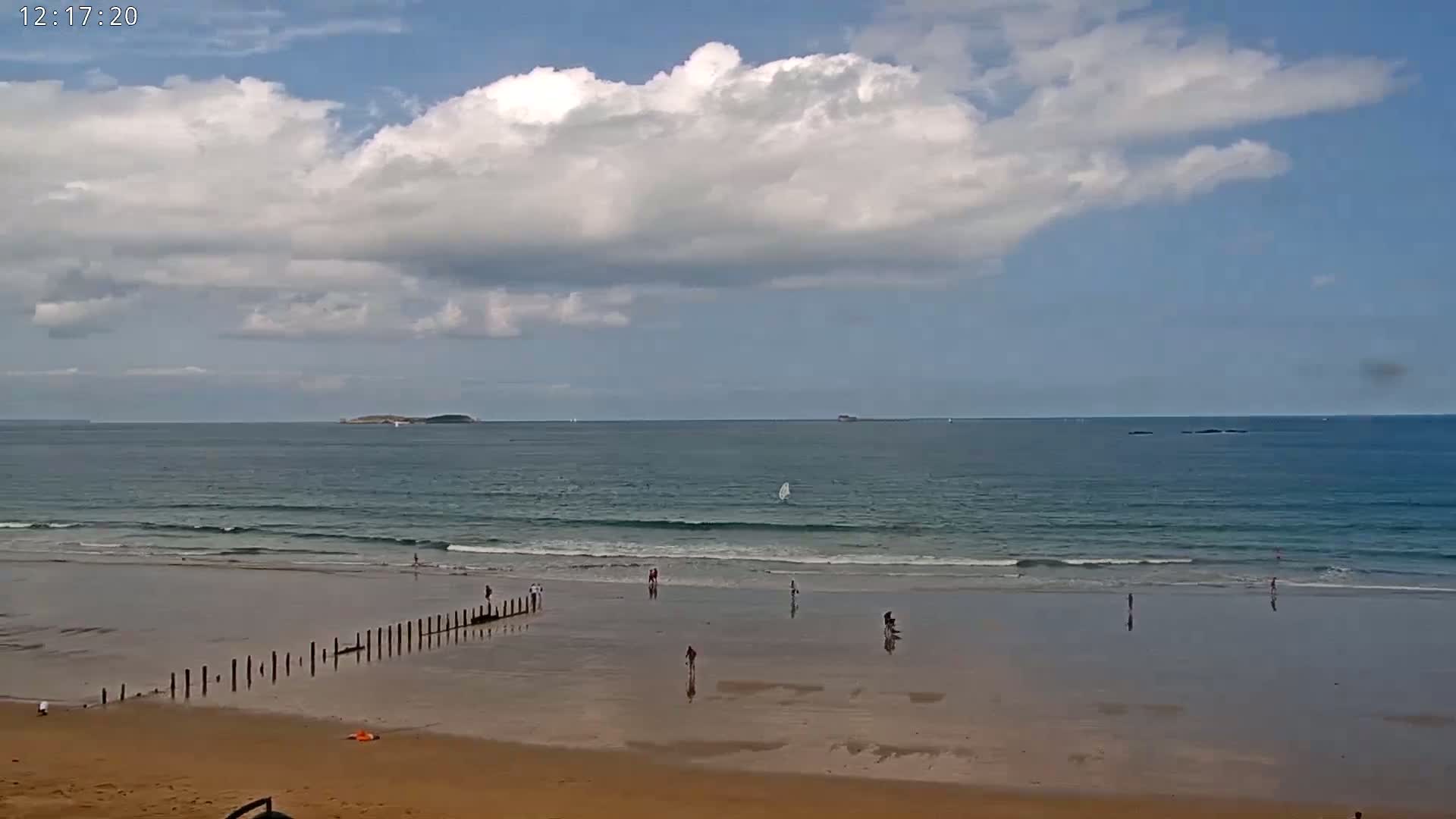 A sandy beach with a few people and a row of posts extends into the ocean under a partly cloudy sky.