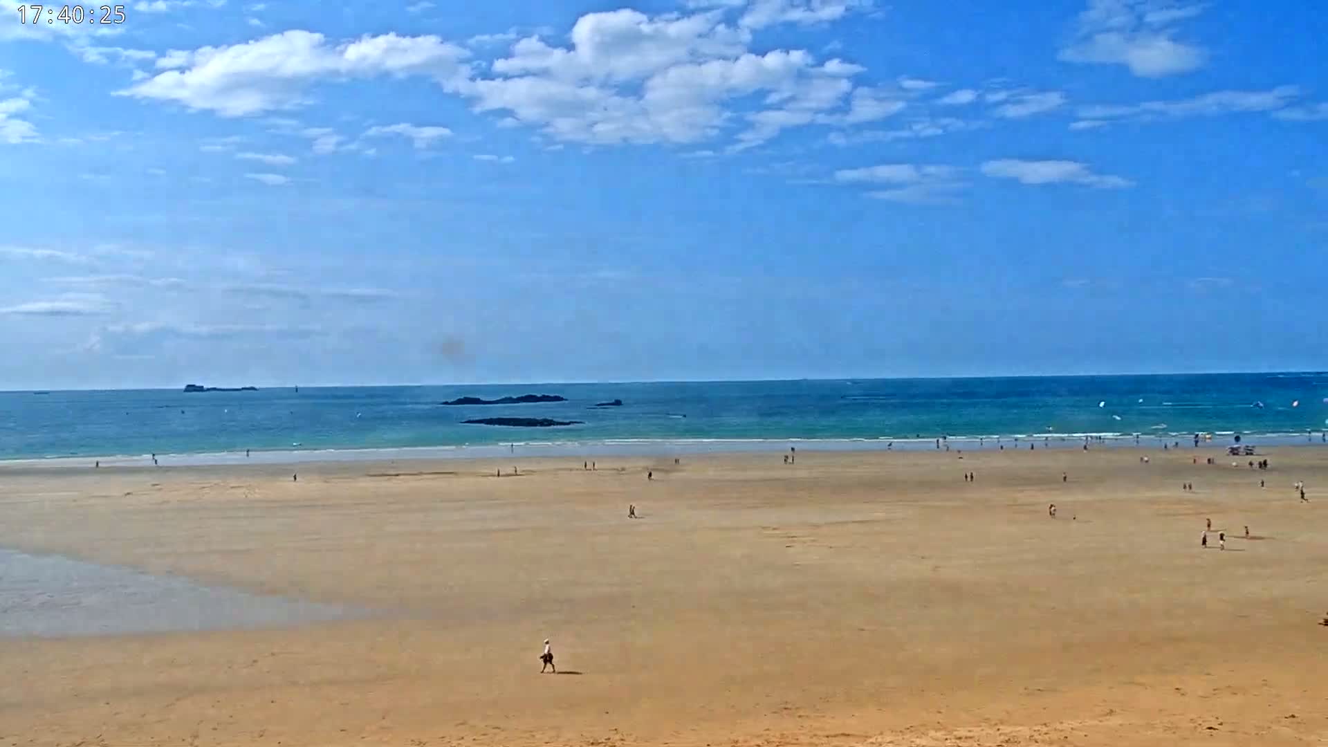 A wide sandy beach stretches to a calm turquoise ocean under a mostly sunny sky with scattered clouds.