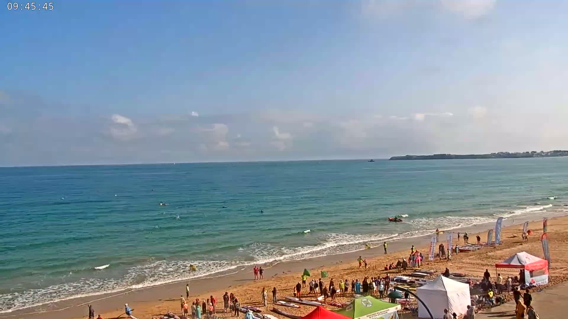 A sandy beach with a calm, clear ocean under a mostly sunny sky shows numerous people gathered near the water's edge, many with what appear to be surfboards or paddleboards.
