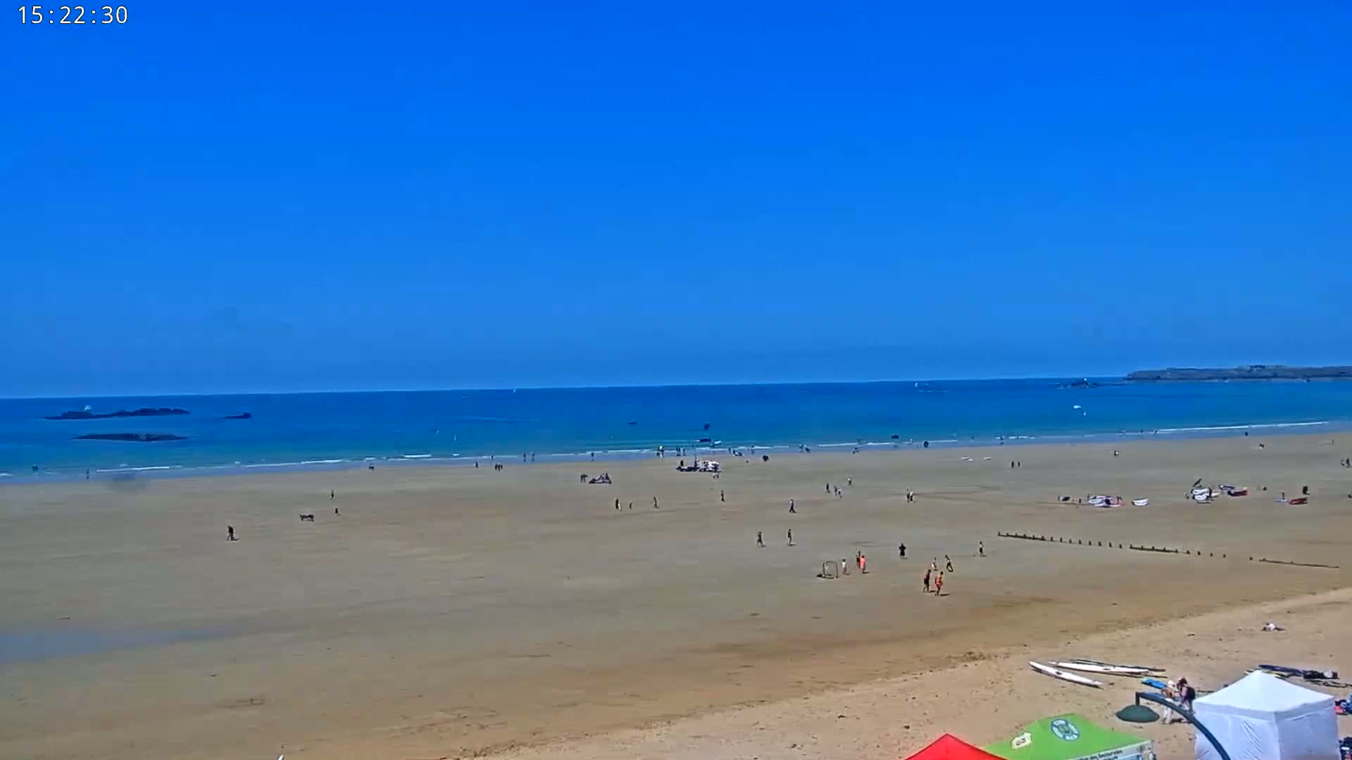 A wide sandy beach with scattered people and some small boats in the ocean under a clear blue sky.