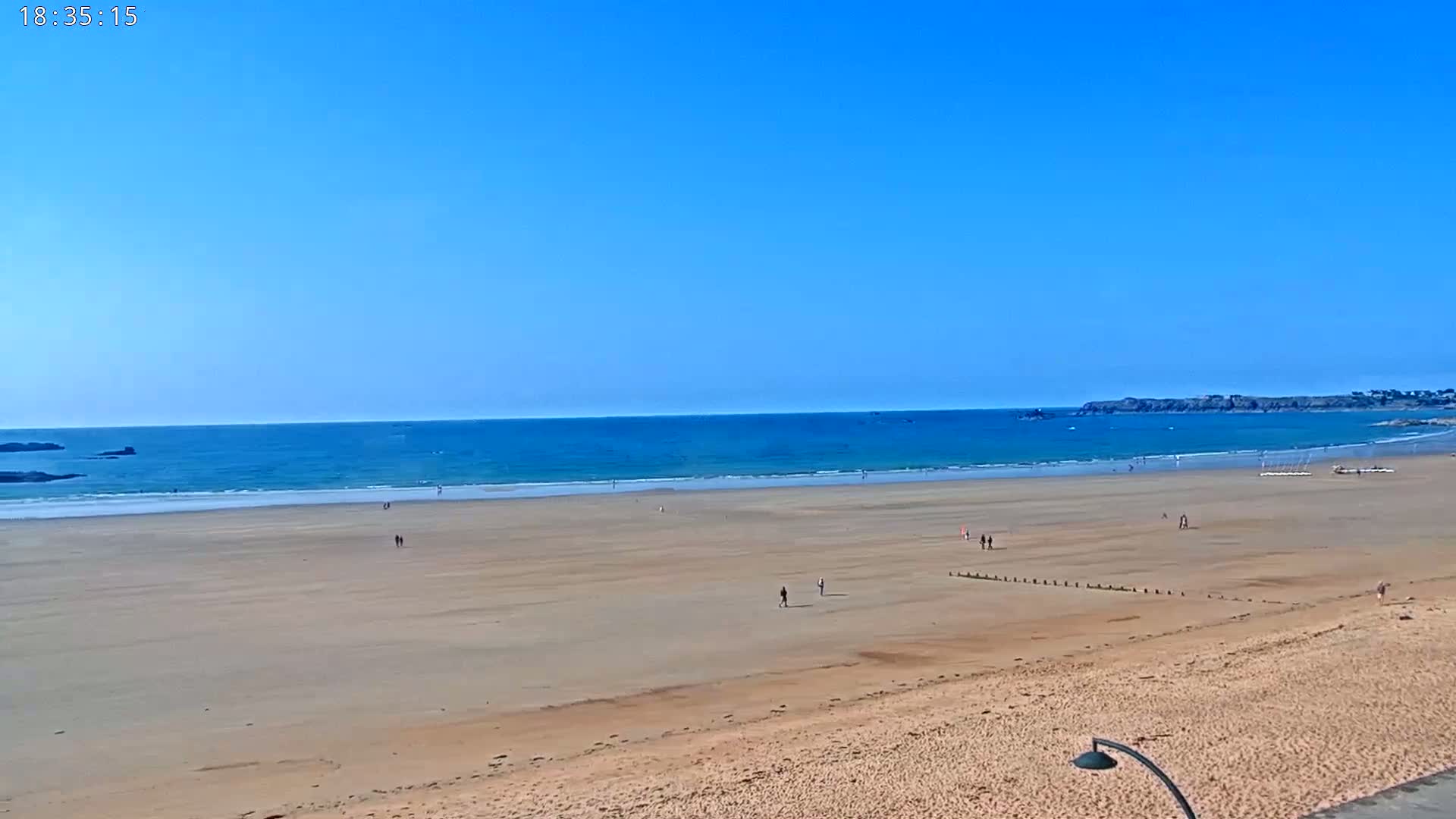 A wide sandy beach stretches to a calm blue ocean under a clear blue sky, with a few people scattered across the sand.