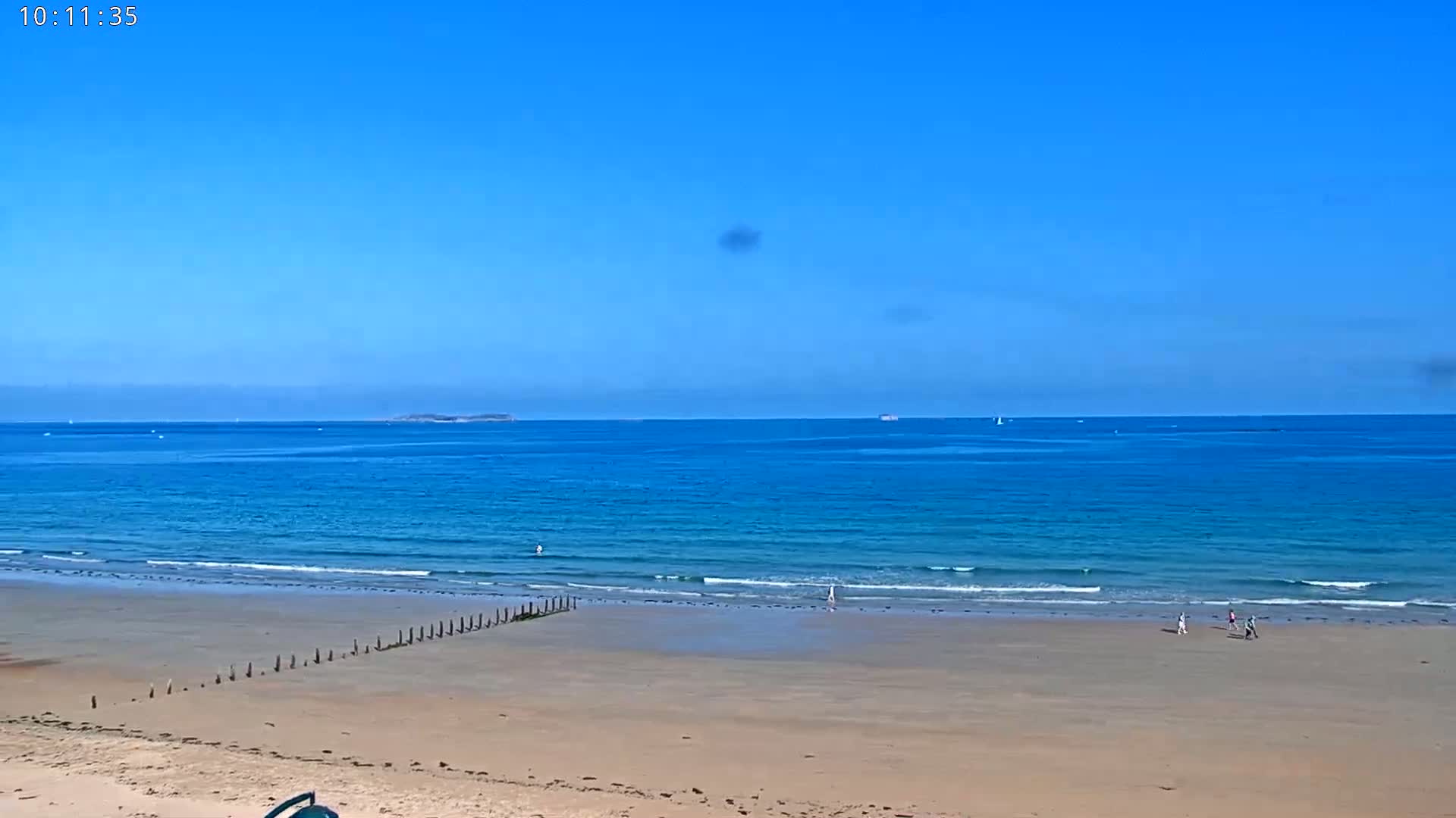 A sandy beach with a few people walking on it stretches to a calm, blue ocean under a clear blue sky.