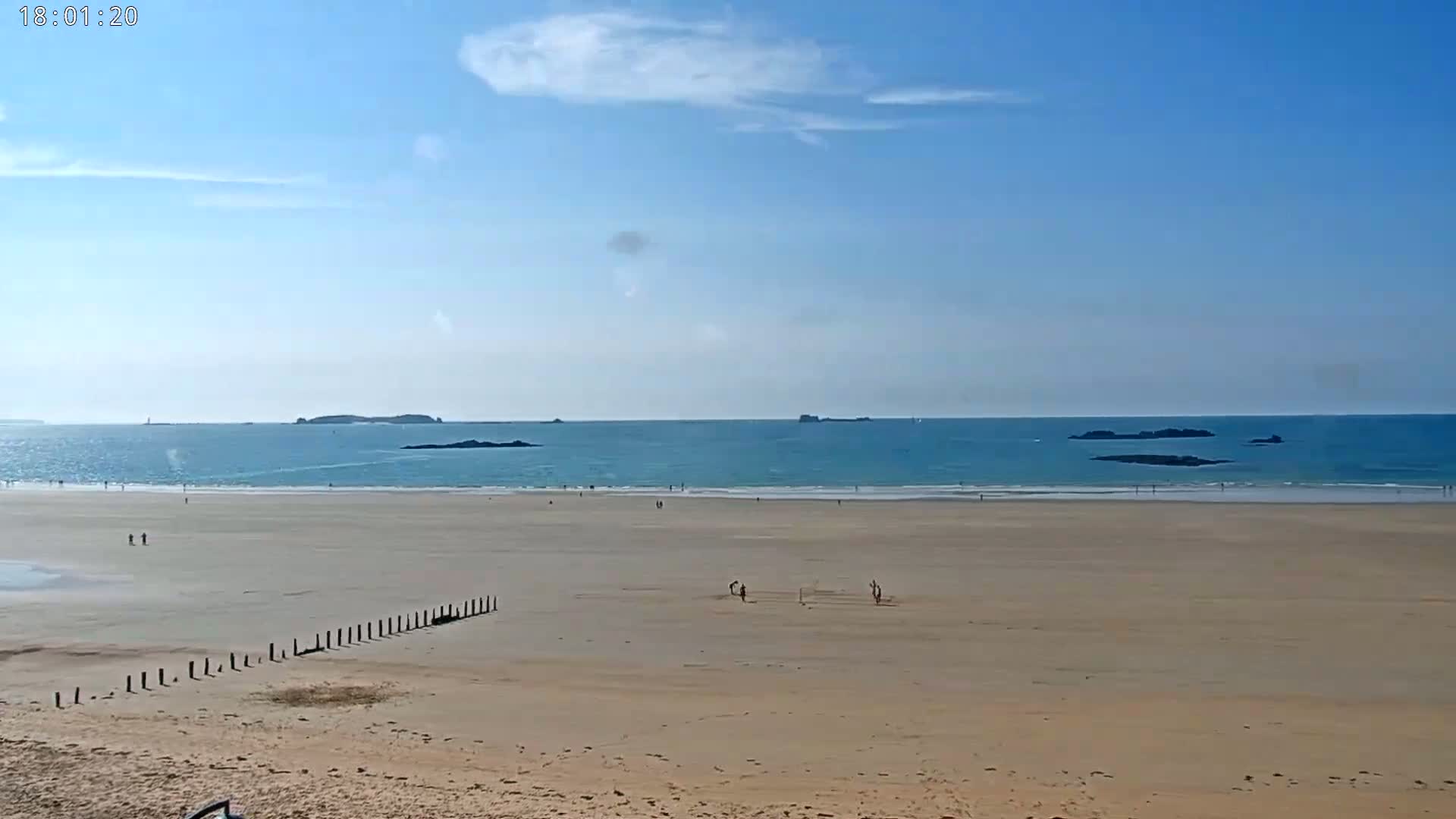 A mostly sunny day at a wide sandy beach shows a calm, light-blue ocean with small islands in the distance and a few people scattered on the sand.