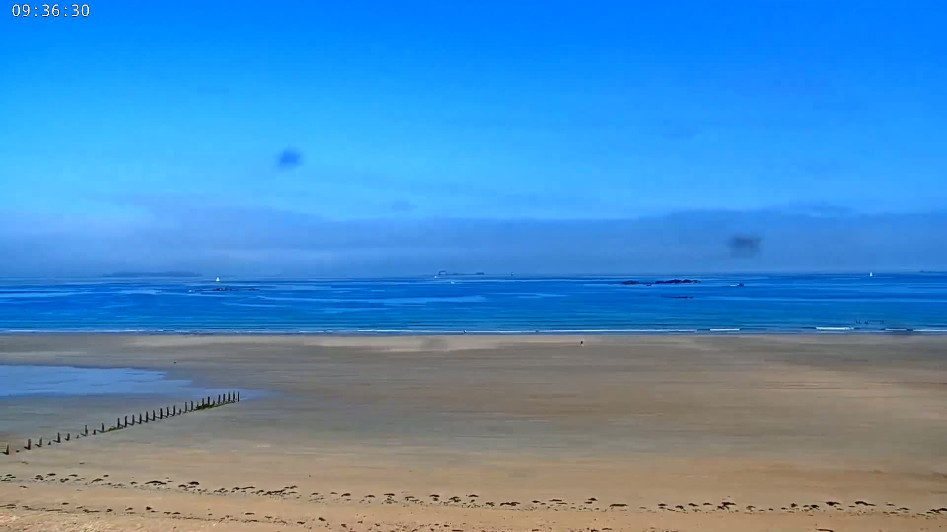 A wide sandy beach stretches to a calm blue ocean under a mostly clear blue sky.
