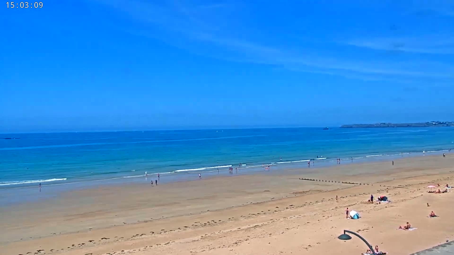 A sandy beach with scattered people stretches to a calm, turquoise ocean under a clear blue sky.